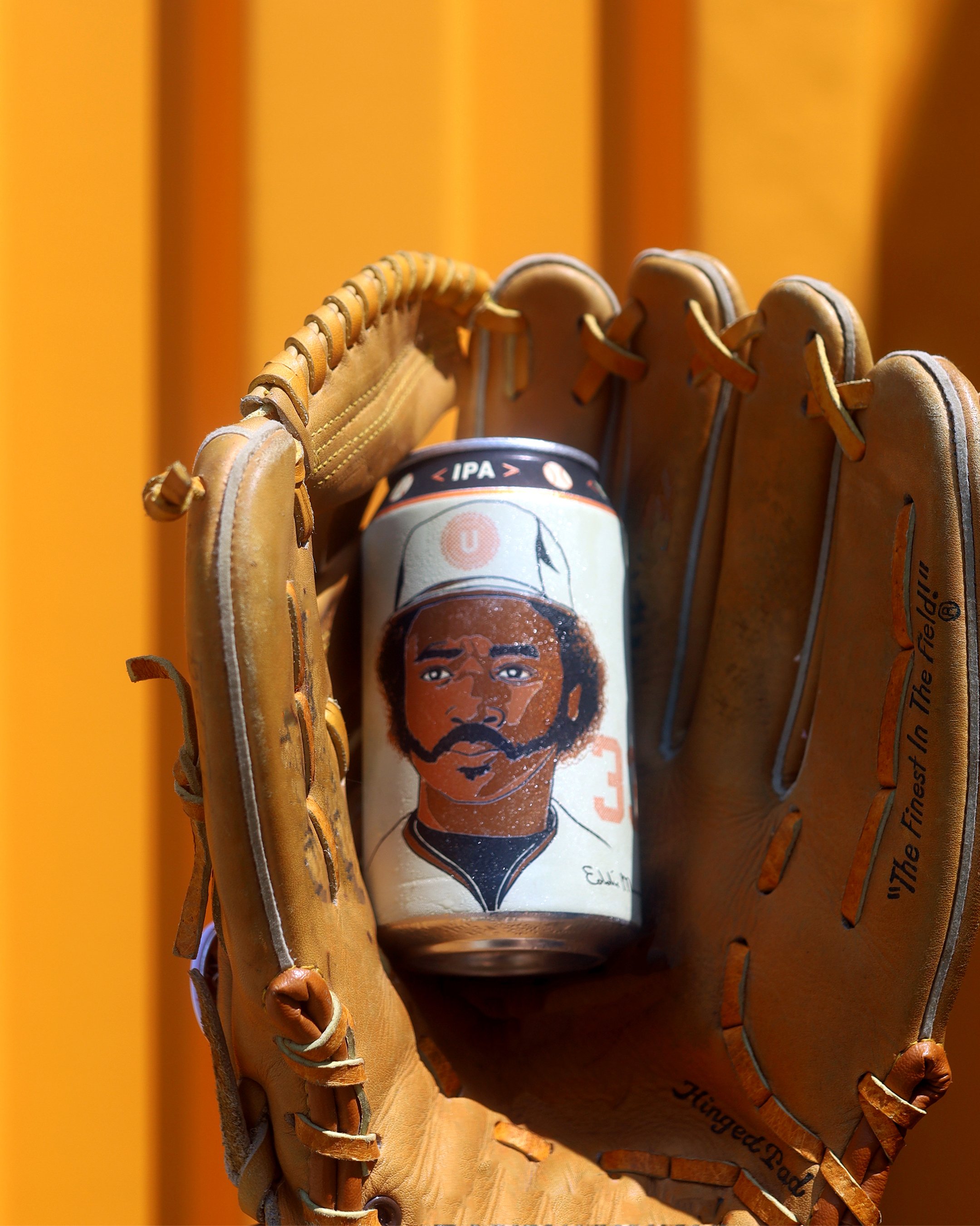 A baseball glove holding a can of IPA beer with a hand-drawn portrait of Eddie Murray