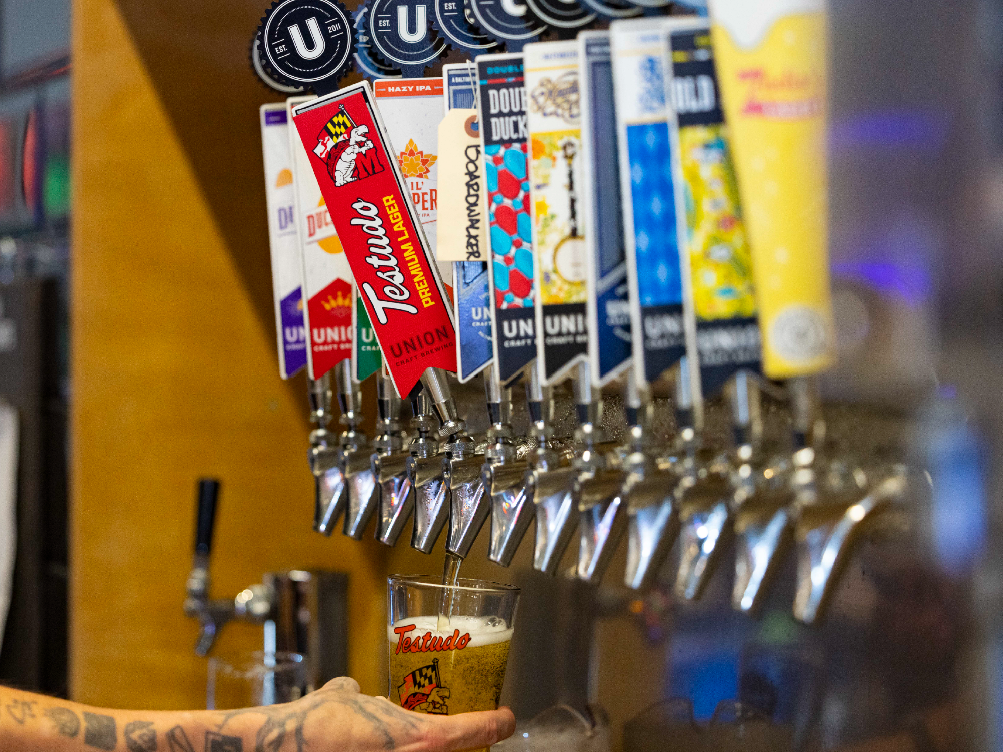 A row of beer taps with colorful tap handles in a bar, as a hand pours a beer into a glass with the Testado logo.