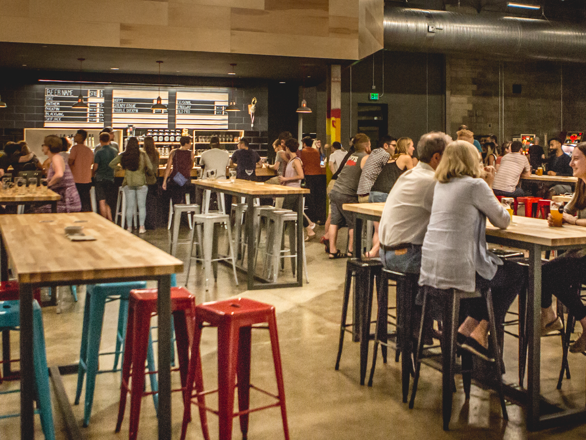 A busy bar with many people sitting and standing at the counter, socializing and drinking. The bar area has a black wall with a menu board and hanging lights. There are tables and stools, some of which are occupied. The setting appears lively and modern.