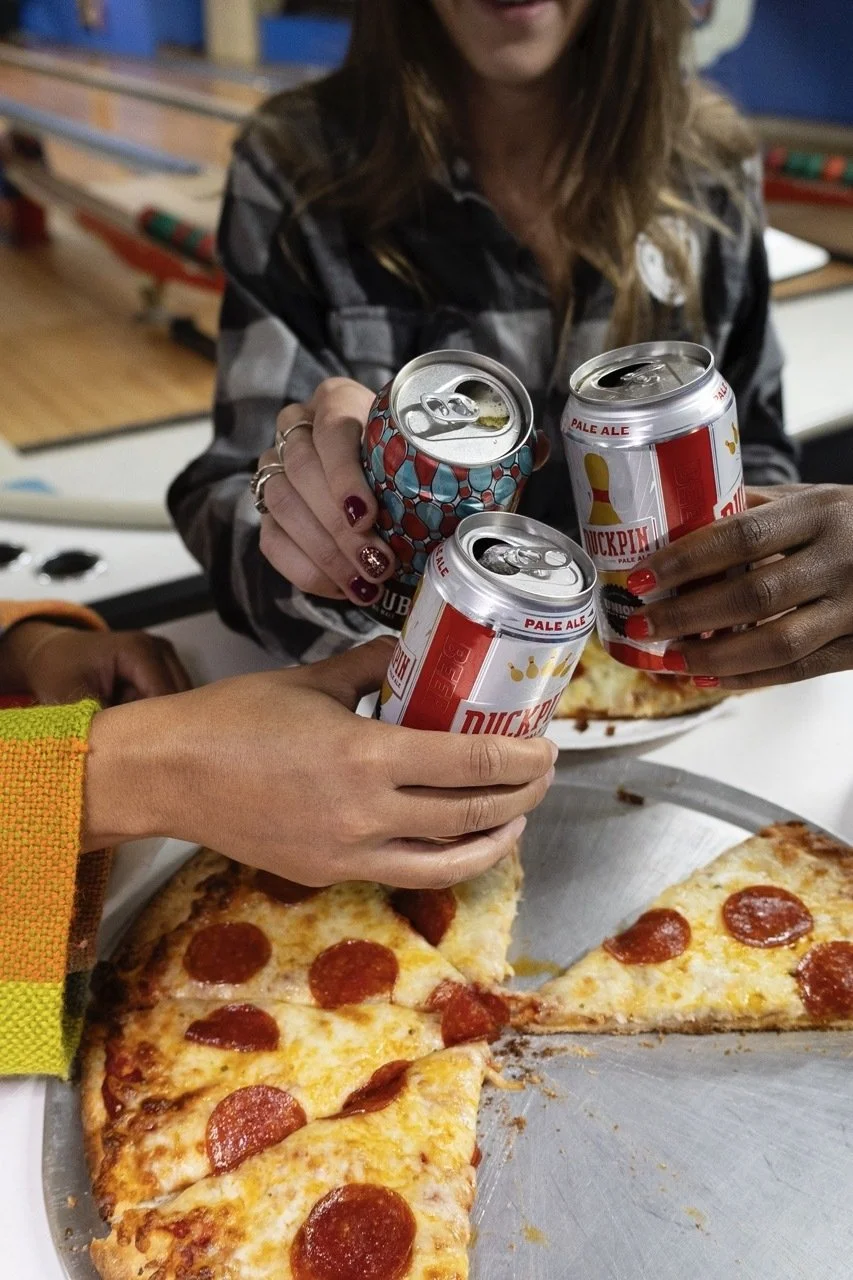Three hands cheersing Duckpin Pale Ale and Double Duckpin at a bowling alley with pizza on table