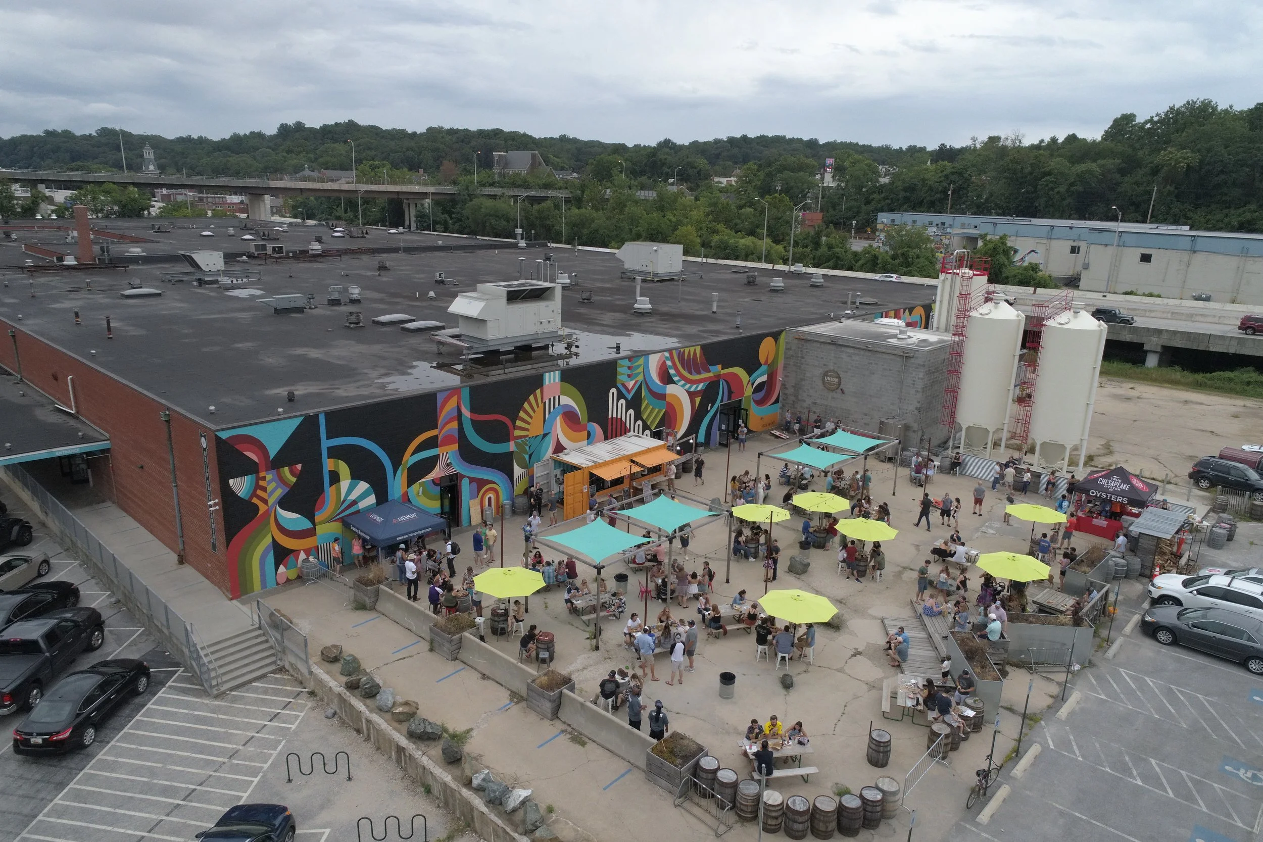 An outdoor patio with people sitting at tables under yellow umbrellas in front of a mural on a building, with parking spaces and cars nearby.