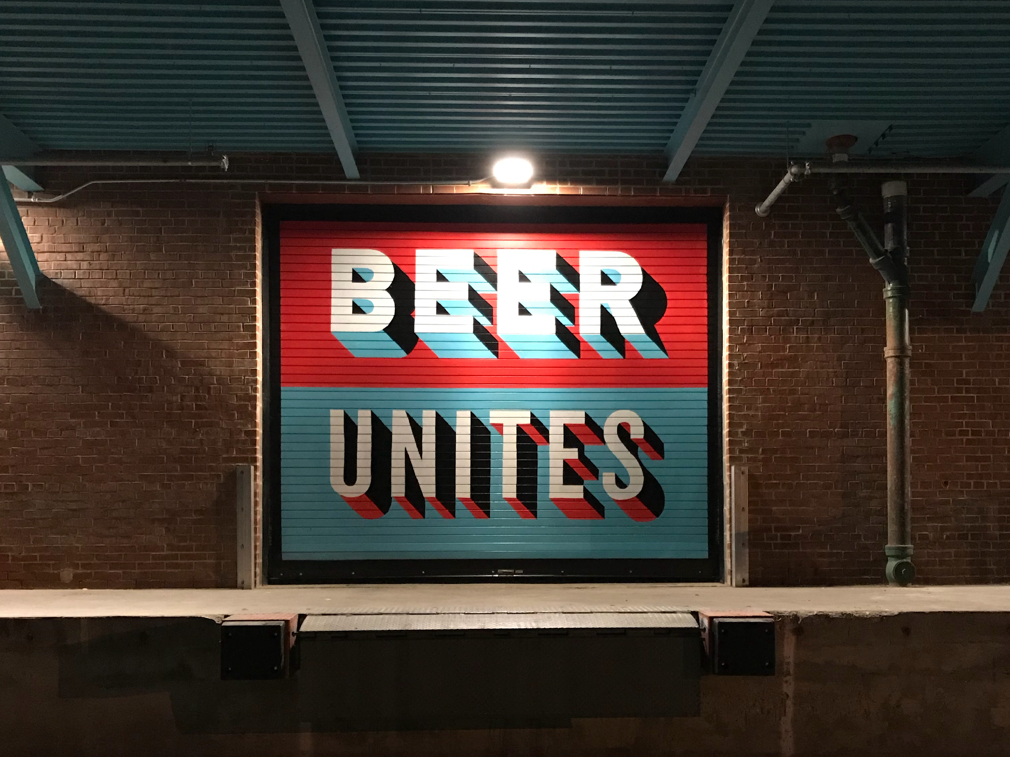 Sign with the words "BEER UNITED" in large, stylized letters, mounted on a brick wall under a blue metal roof.