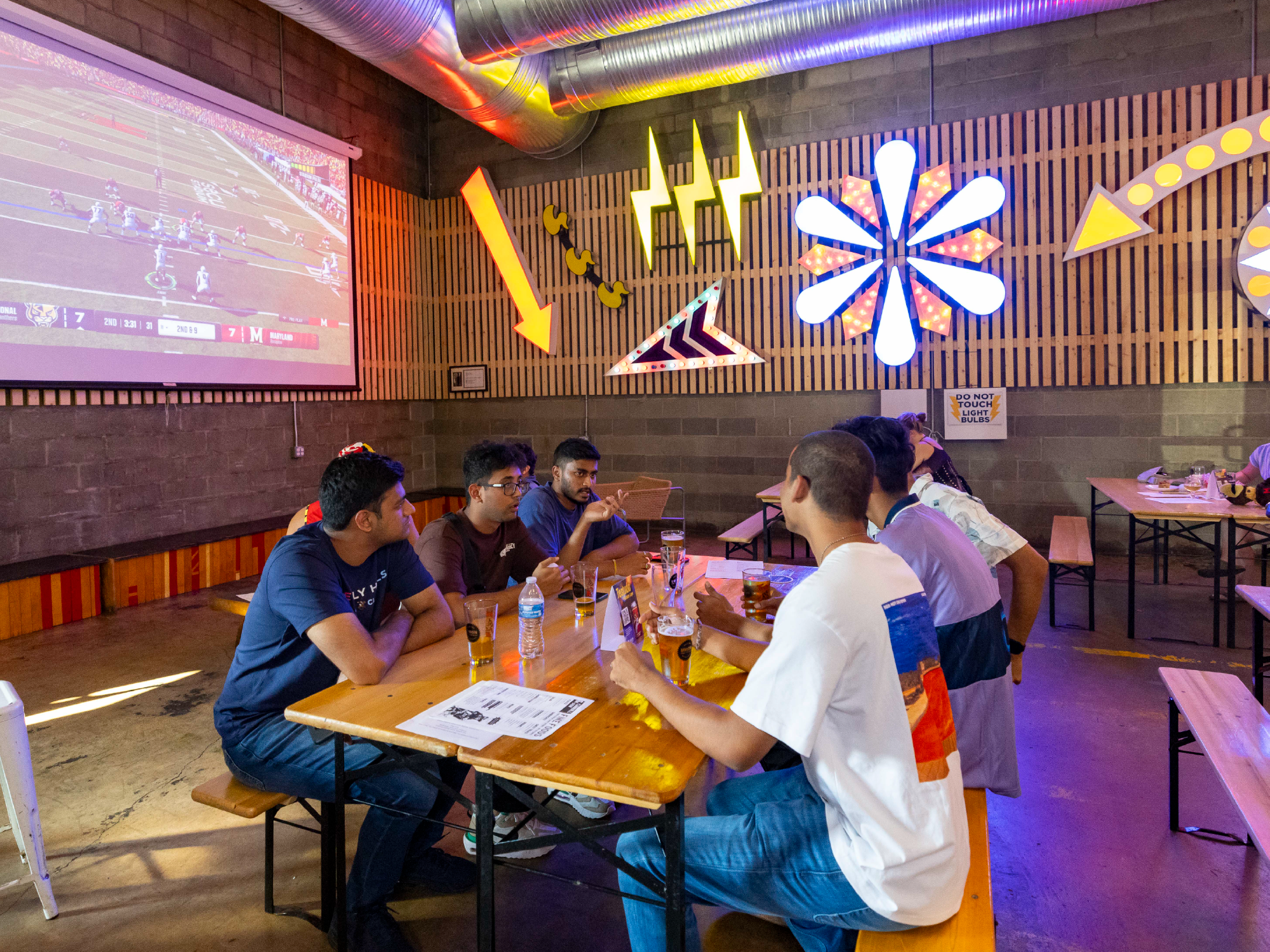 Group of young people watching a football game on a large screen at a bar or restaurant, with vibrant neon signs on the wall.