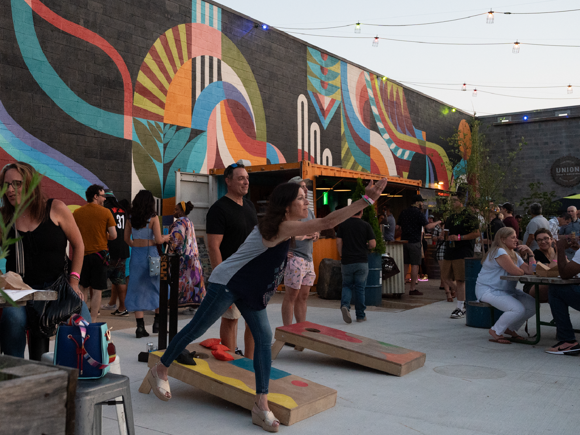 A woman participating in a game of cornhole at an outdoor event during the evening, with a crowd of people socializing and a colorful mural on the building wall in the background.