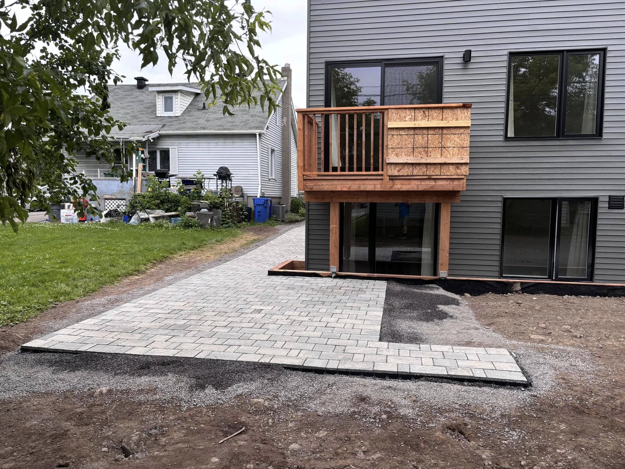 Two houses with a newly constructed deck and patio, featuring gray siding and windows. A freshly laid stone pathway leads to the back entrance area. The yard has construction materials and a grassy area, with a neighboring house visible in the backgr