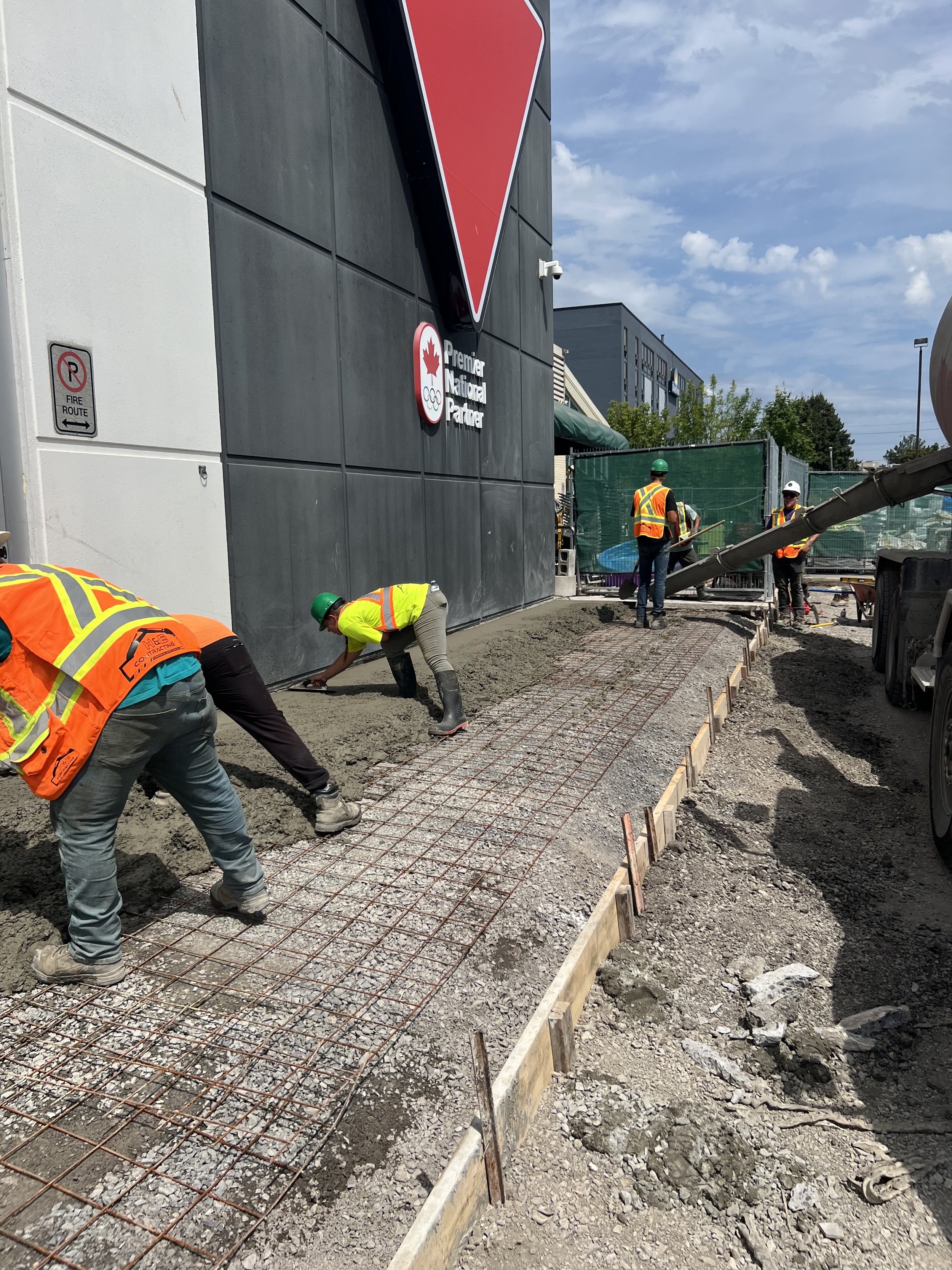Construction workers pouring concrete on a wire-reinforced path next to a building with a triangular logo.