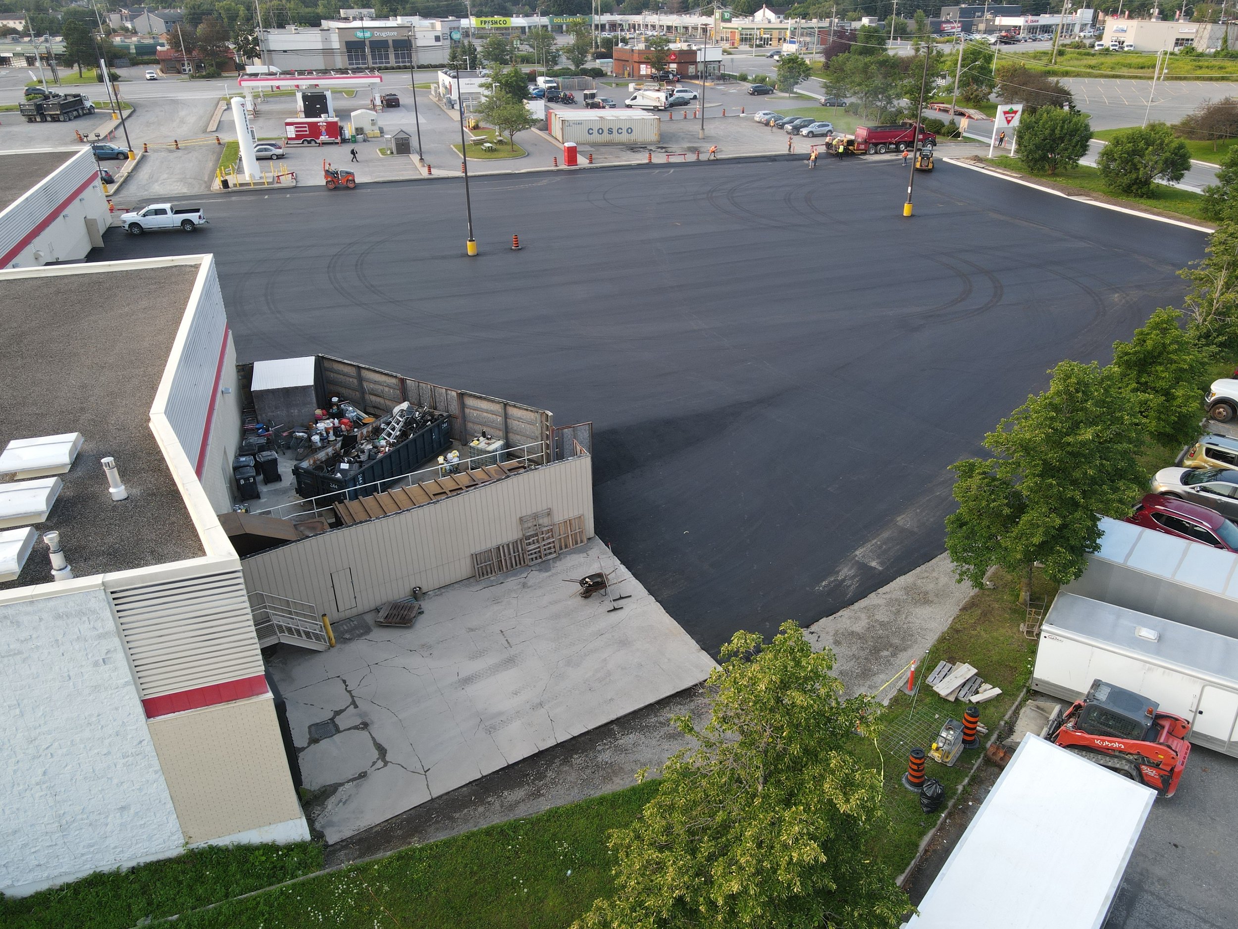 An aerial view of a large parking lot with freshly paved asphalt, surrounded by buildings and trees. A few vehicles and construction equipment are visible. There is a fenced area at the corner of a building with assorted items and a dumpster. Nearby