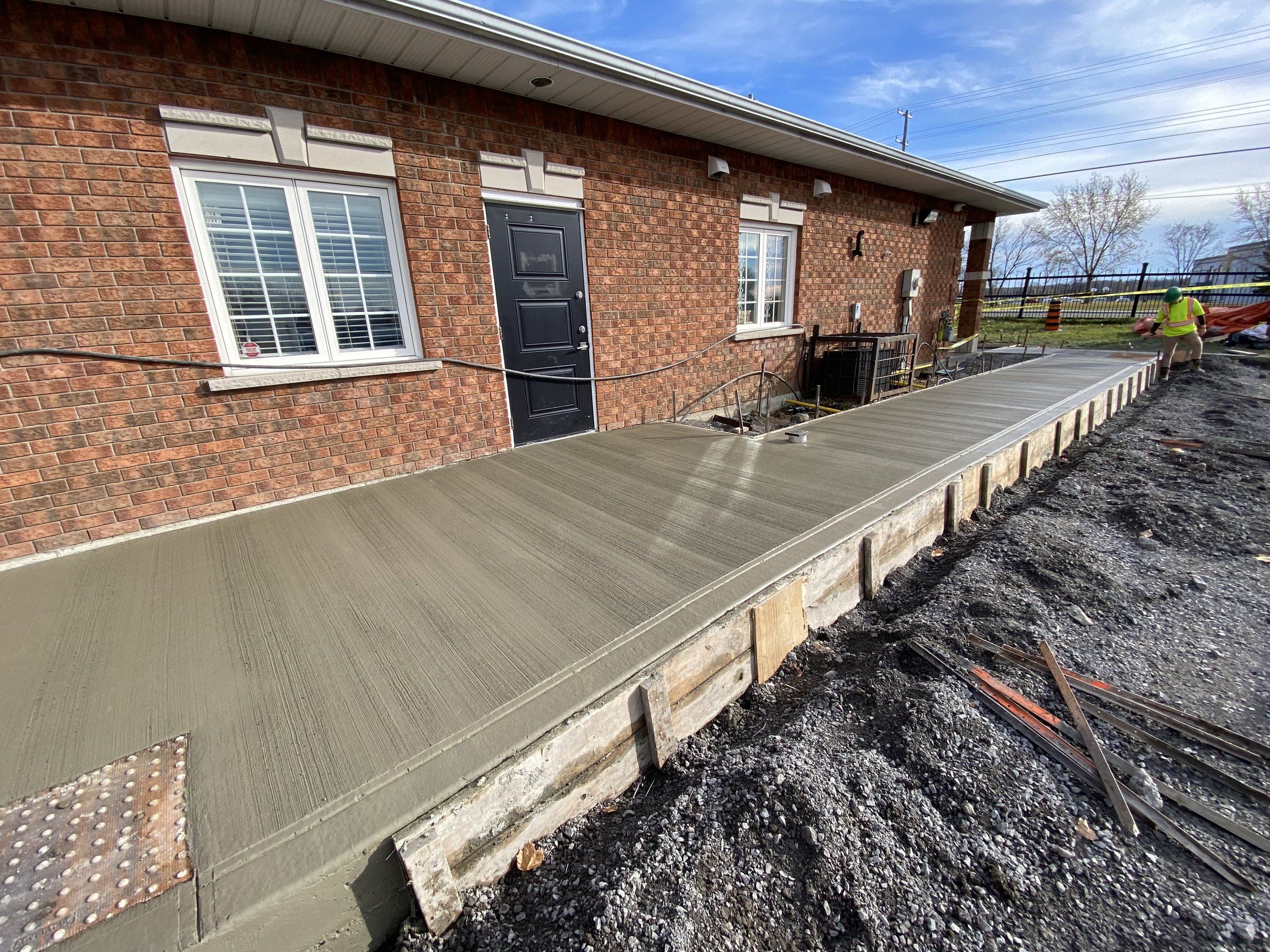 Freshly poured concrete ramp next to a brick building with windows and a black door, construction tools and materials visible, indicating an ongoing construction project.