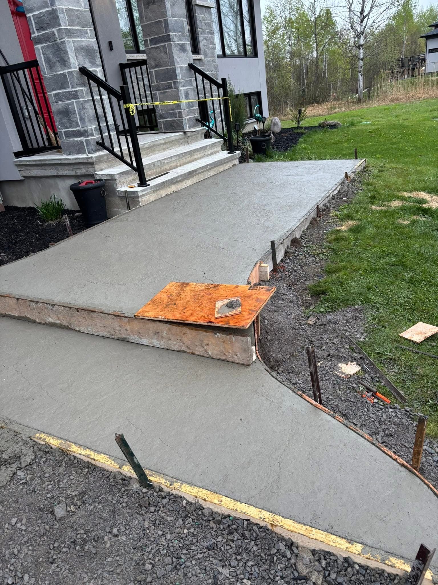 Freshly poured concrete walkway leading to a house entrance with black railings and stone facade.
