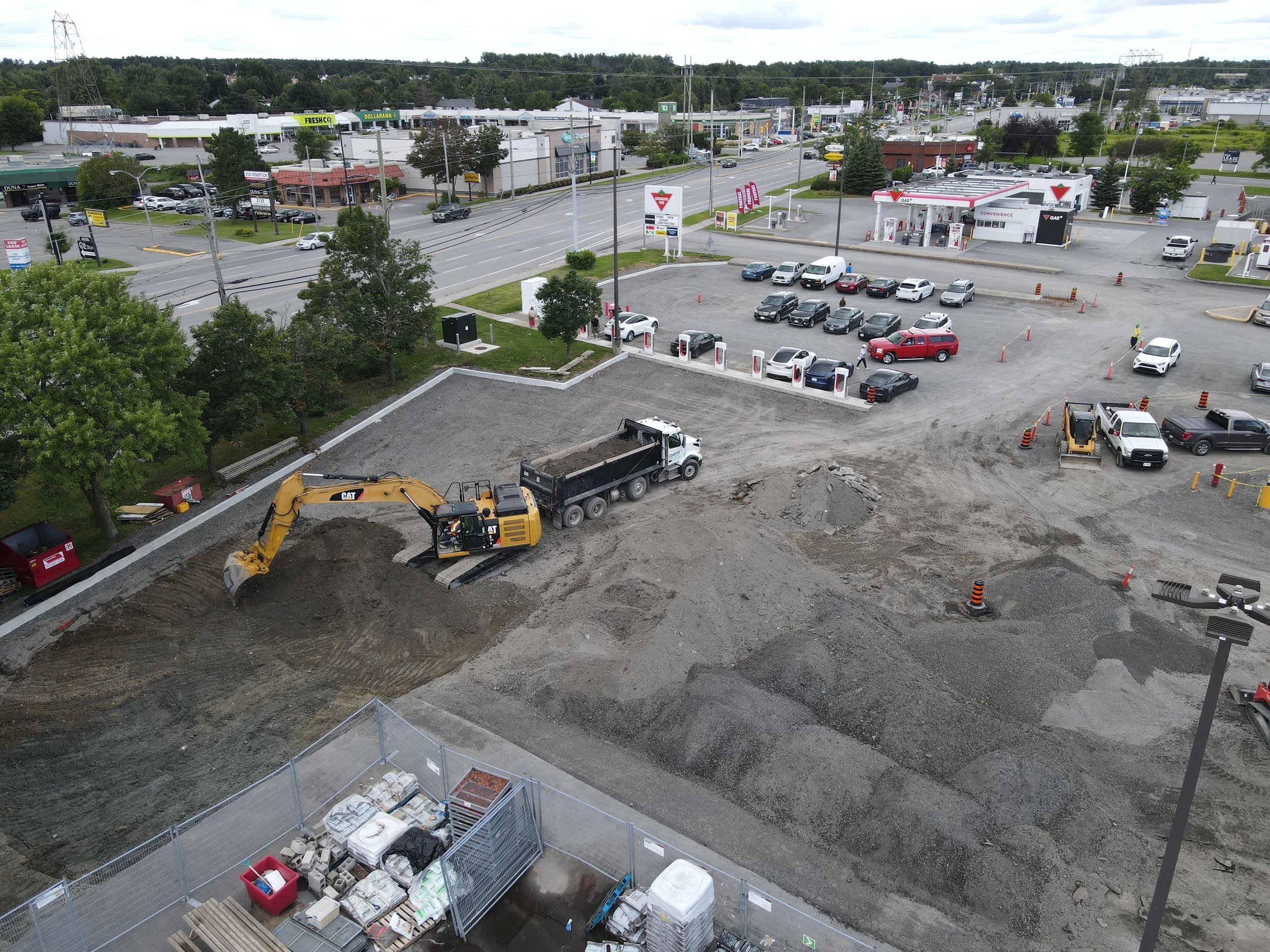 Aerial view of a construction site near a gas station, showing an excavator and dump truck working on the site. There are piles of gravel and equipment, with a parking lot and vehicles nearby.