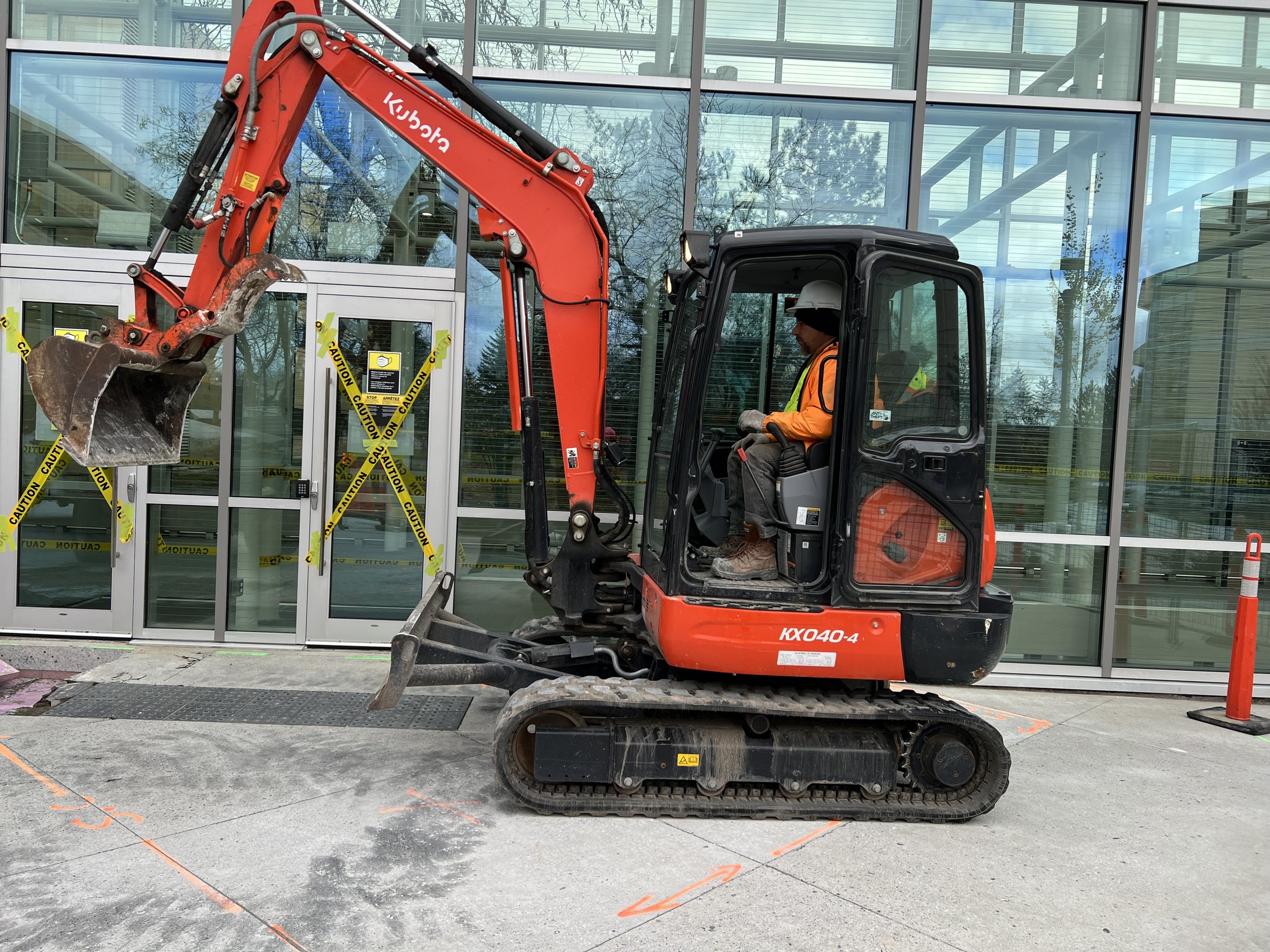 A red mini excavator with a worker in an orange safety vest is parked in front of a building entrance marked with caution tape.