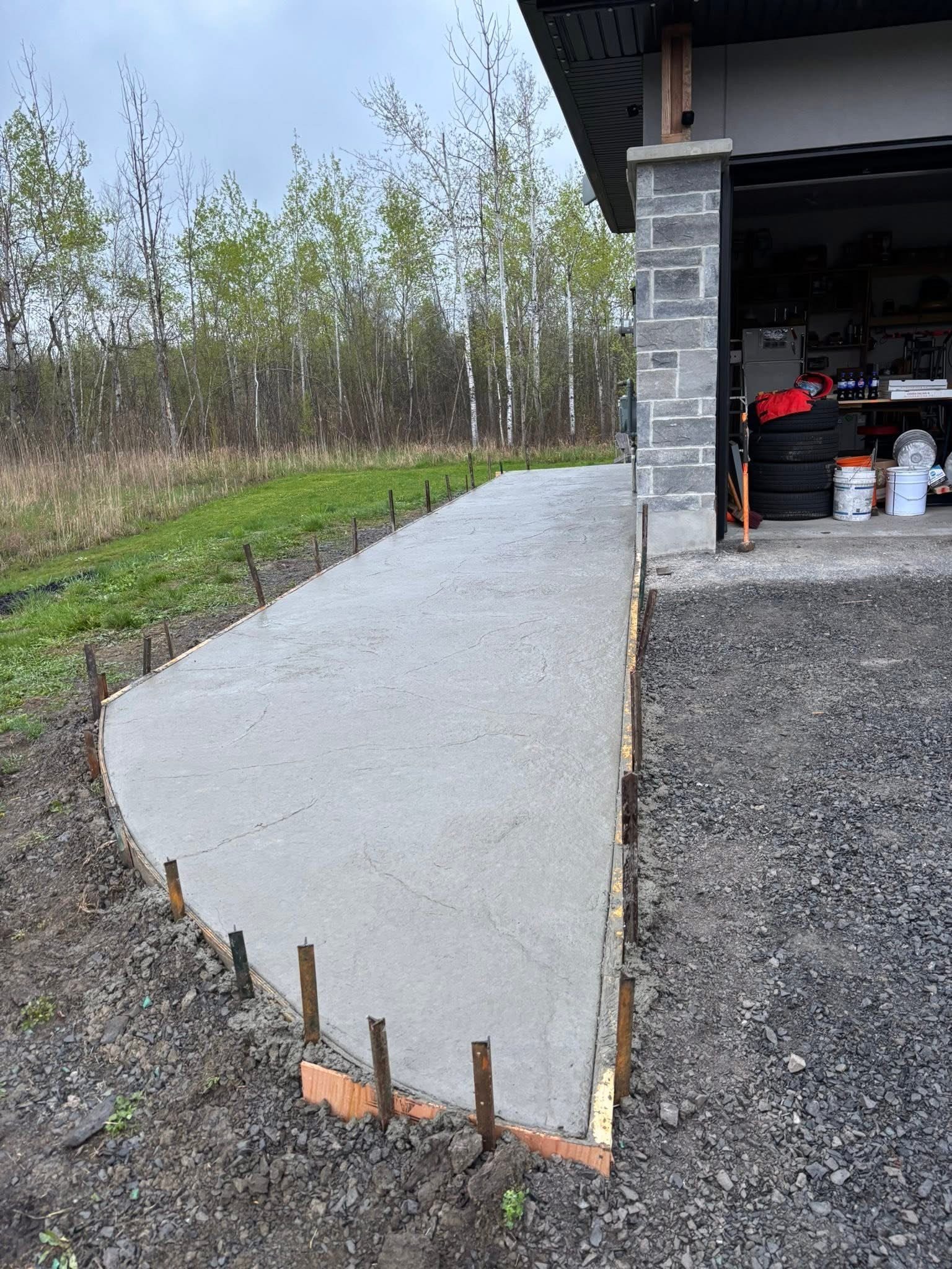 Freshly poured concrete driveway next to a garage with tools and equipment inside, surrounded by trees and grass.