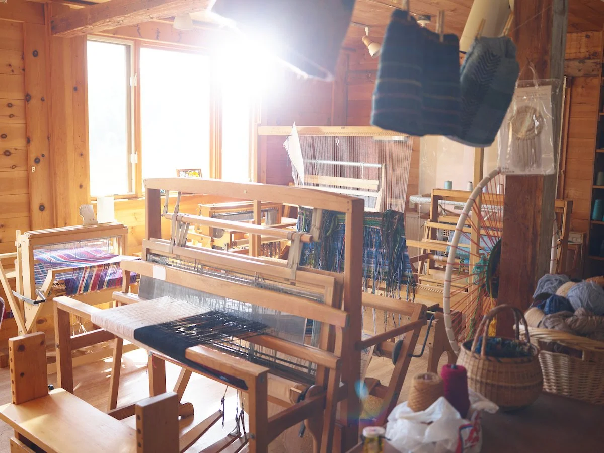 A cozy wooden craft room with sunlight streaming through a large window, filled with weaving looms and baskets of yarn and fabric.