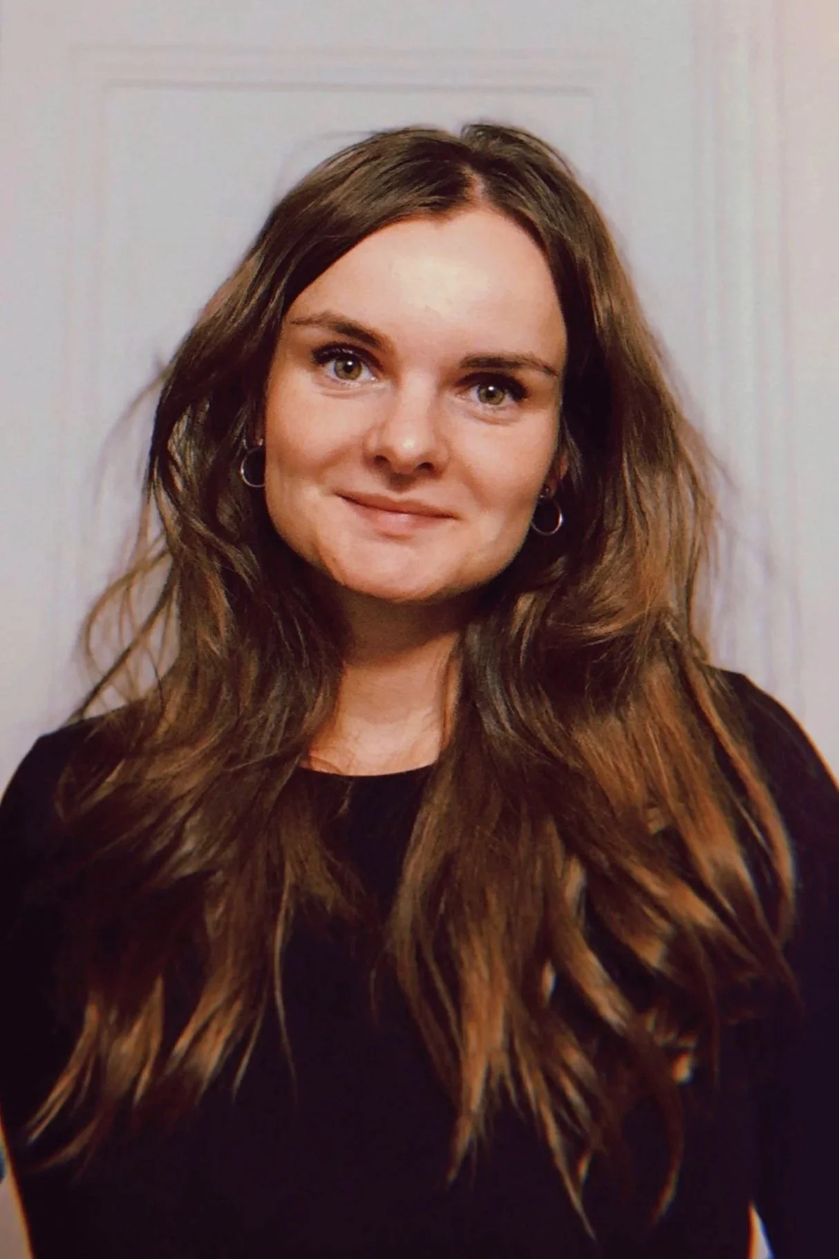 Jeune femme avec longs cheveux bruns, portant un sweatshirt noir, souriant légèrement, portant des boucles d'oreilles en cercles, debout contre un mur blanc.