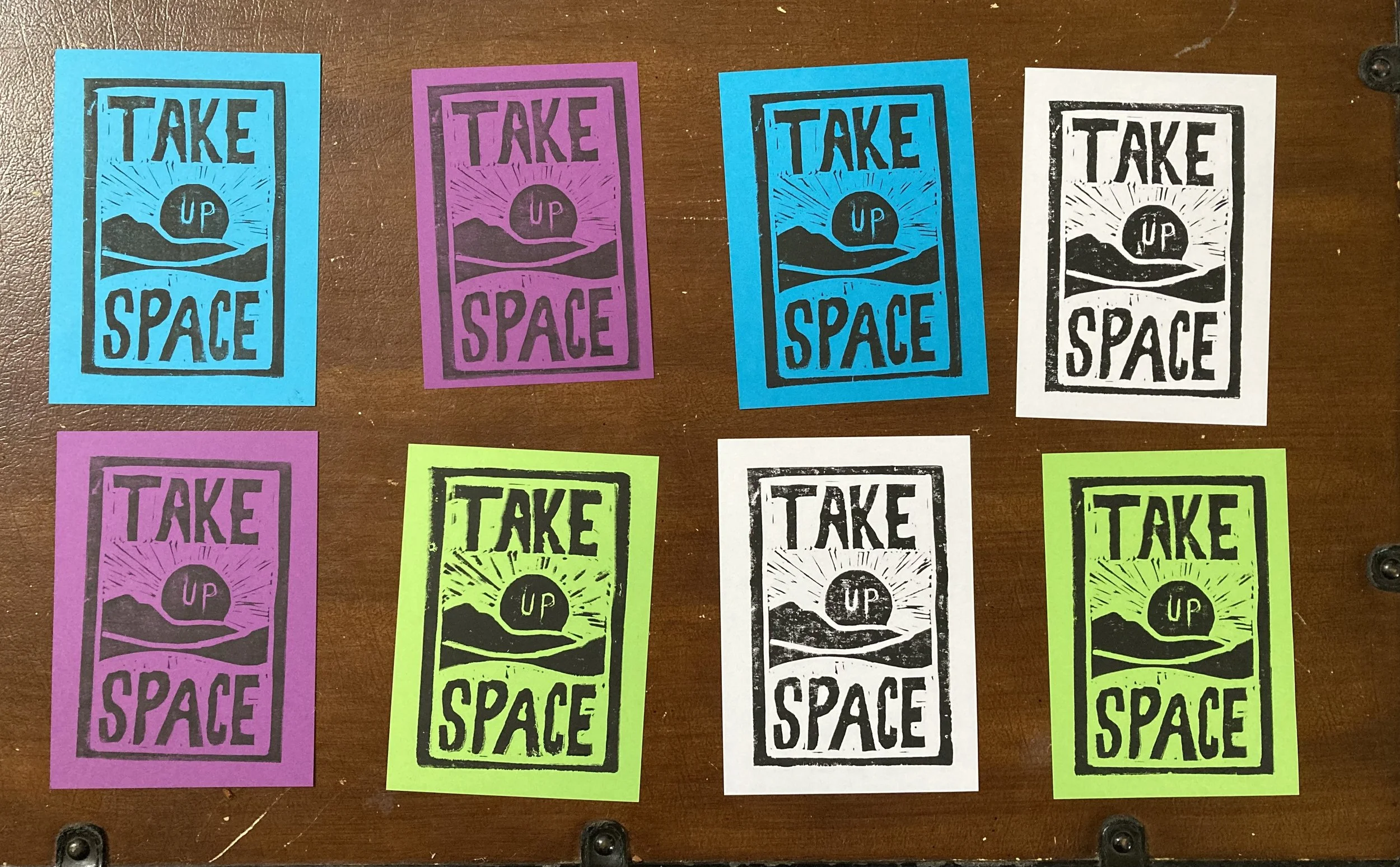 Colorful printed posters with the phrase 'Take Up Space' arranged on a brown wooden surface.
