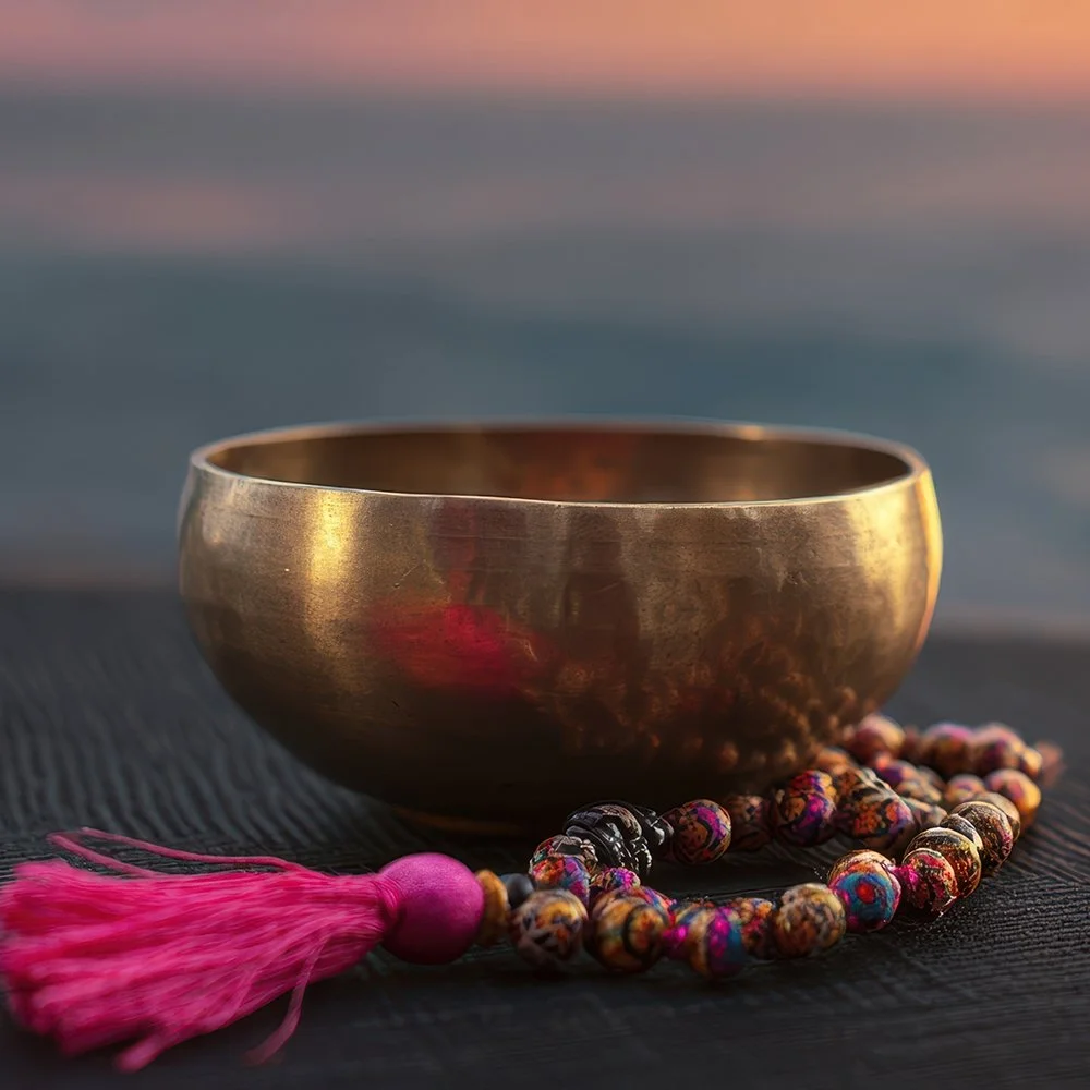 Bol chantant en métal doré avec un mala de prière coloré à côté, posé sur une surface noire avec un fond de coucher de soleil ou de mer calme.