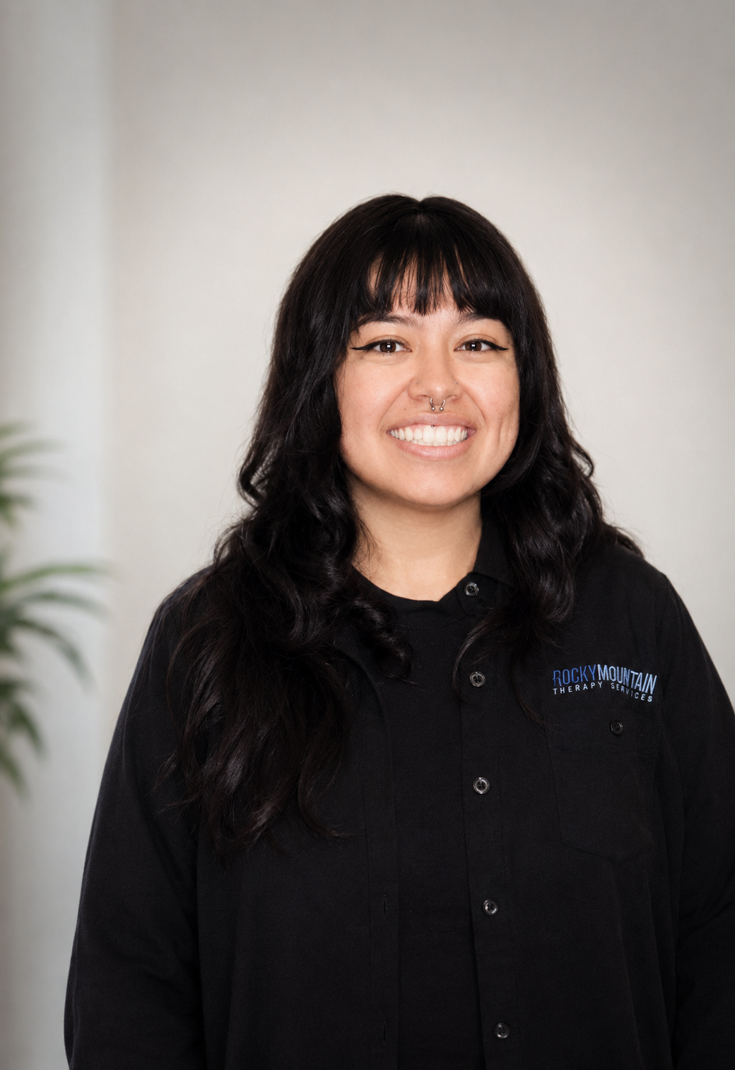 Woman with dark wavy hair, smiling, wearing a black shirt with a logo that says 'Rocky Mountain Therapy'.