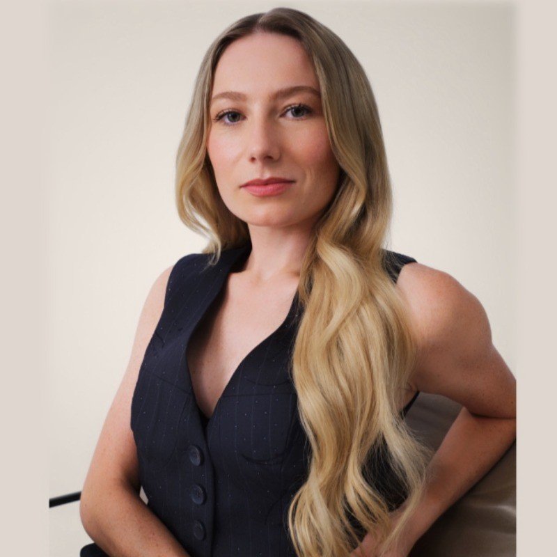A woman sits facing the camera with a confident expression, wearing a dark sleeveless vest, with long blonde hair styled over one shoulder against a neutral background.