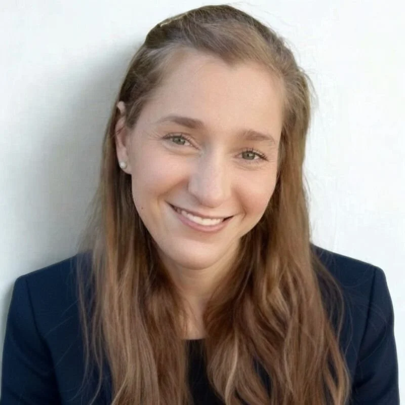 A woman with long dark blonde hair smiles at the camera while wearing a dark blue blazer and small pearl stud earrings sits against a plain white background.