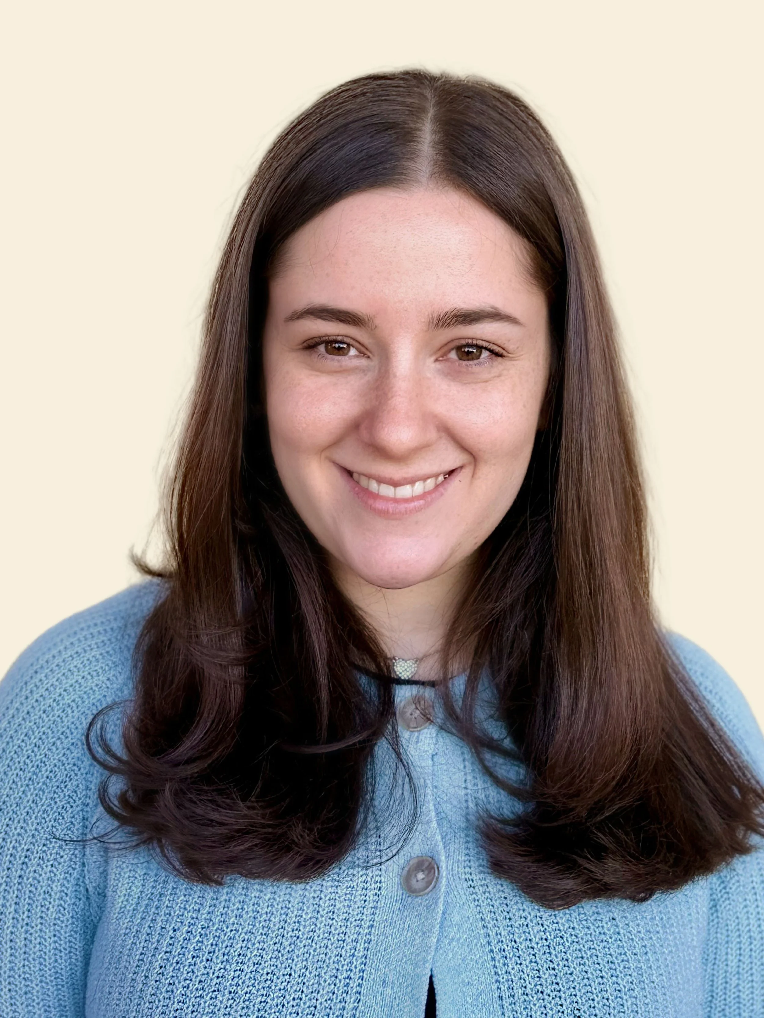 A woman with long brown hair smiles at the camera against a neutral beige background, wearing a light blue sweater.