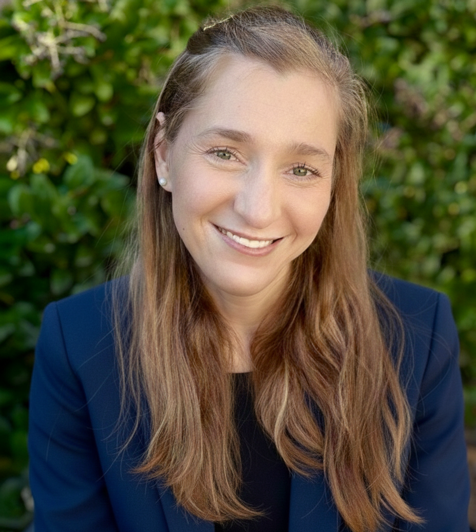 A woman smiling warmly at the camera outdoors wearing a navy blazer with long light brown hair styled half up with greenery in the background.