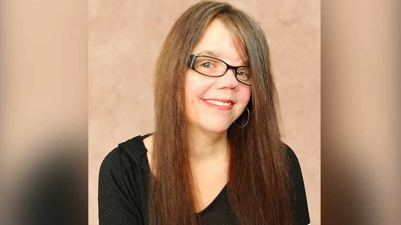 A woman with long brown hair, wearing glasses and hoop earrings, smiling, background is beige.