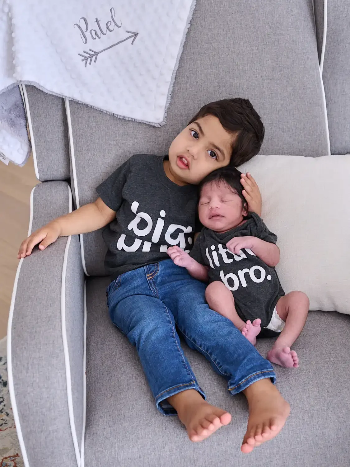 A young boy holding a newborn baby girl while sitting on a gray couch. They are both wearing matching gray T-shirts that say 'big bro.' and 'littlest bro.'
