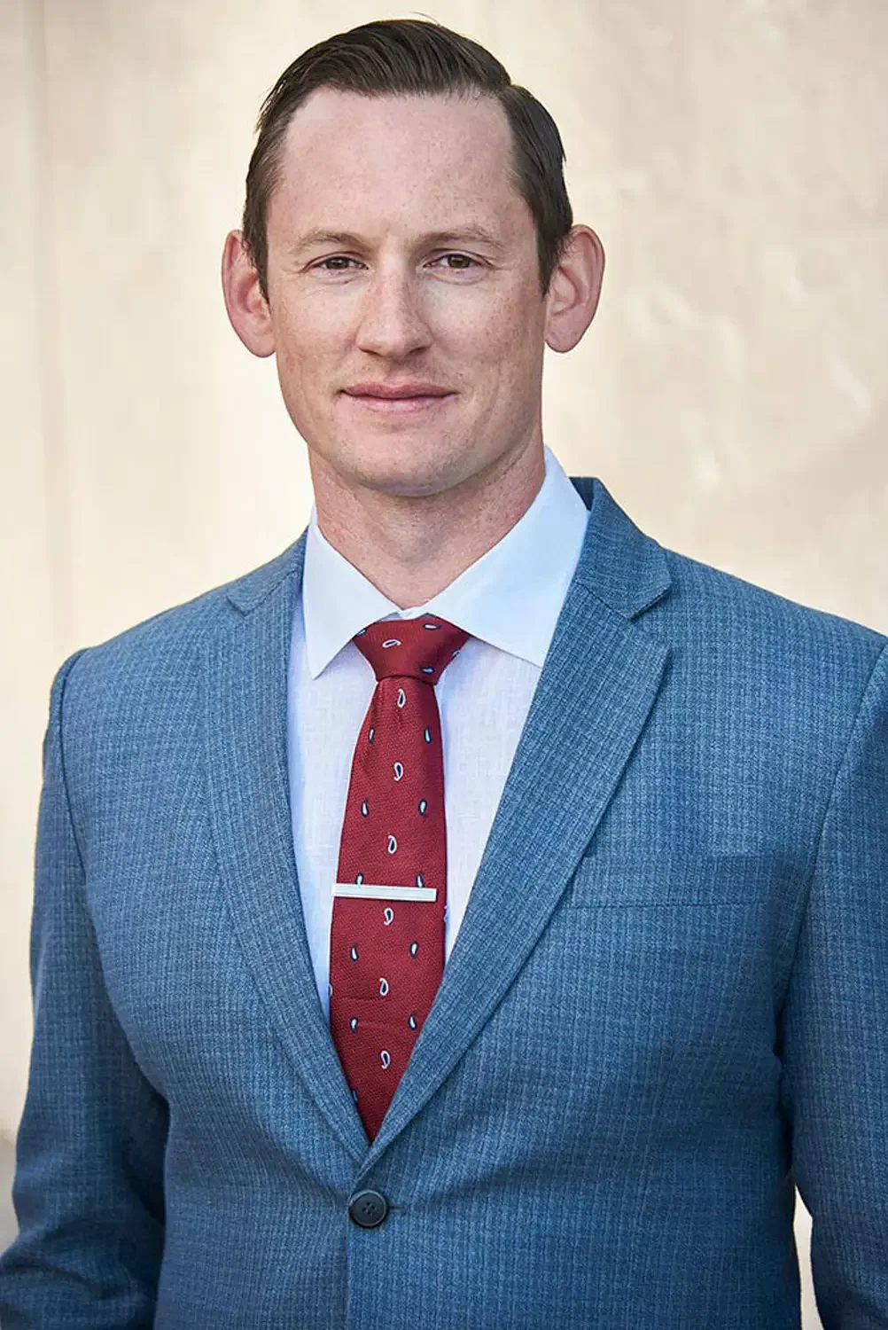 Professional headshot of a young man in a blue suit with a white shirt and red tie, standing outdoors against a neutral background, with a slight smile.