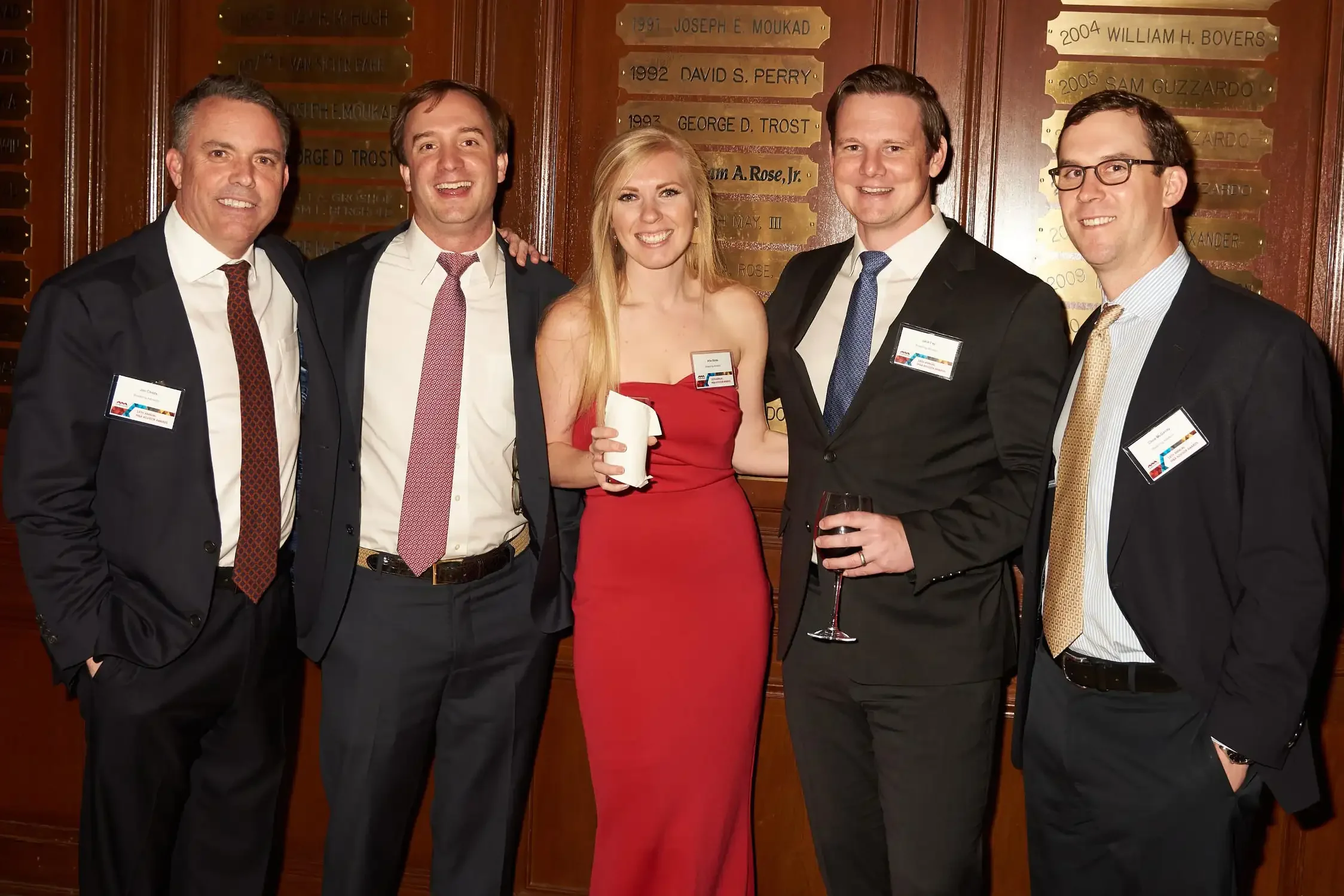 A group of five people dressed in formal attire, standing close together and smiling for the camera in a wood-paneled room with commemorative plaques on the wall behind them.