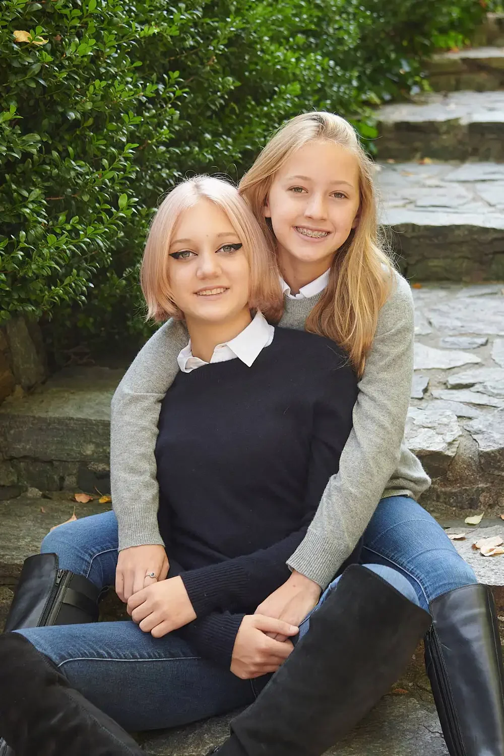 Photo of two young sister's sitting on stone steps outdoors, smiling at the camera. They are dressed casually, with one wearing a black sweater and the other in a gray sweater.
