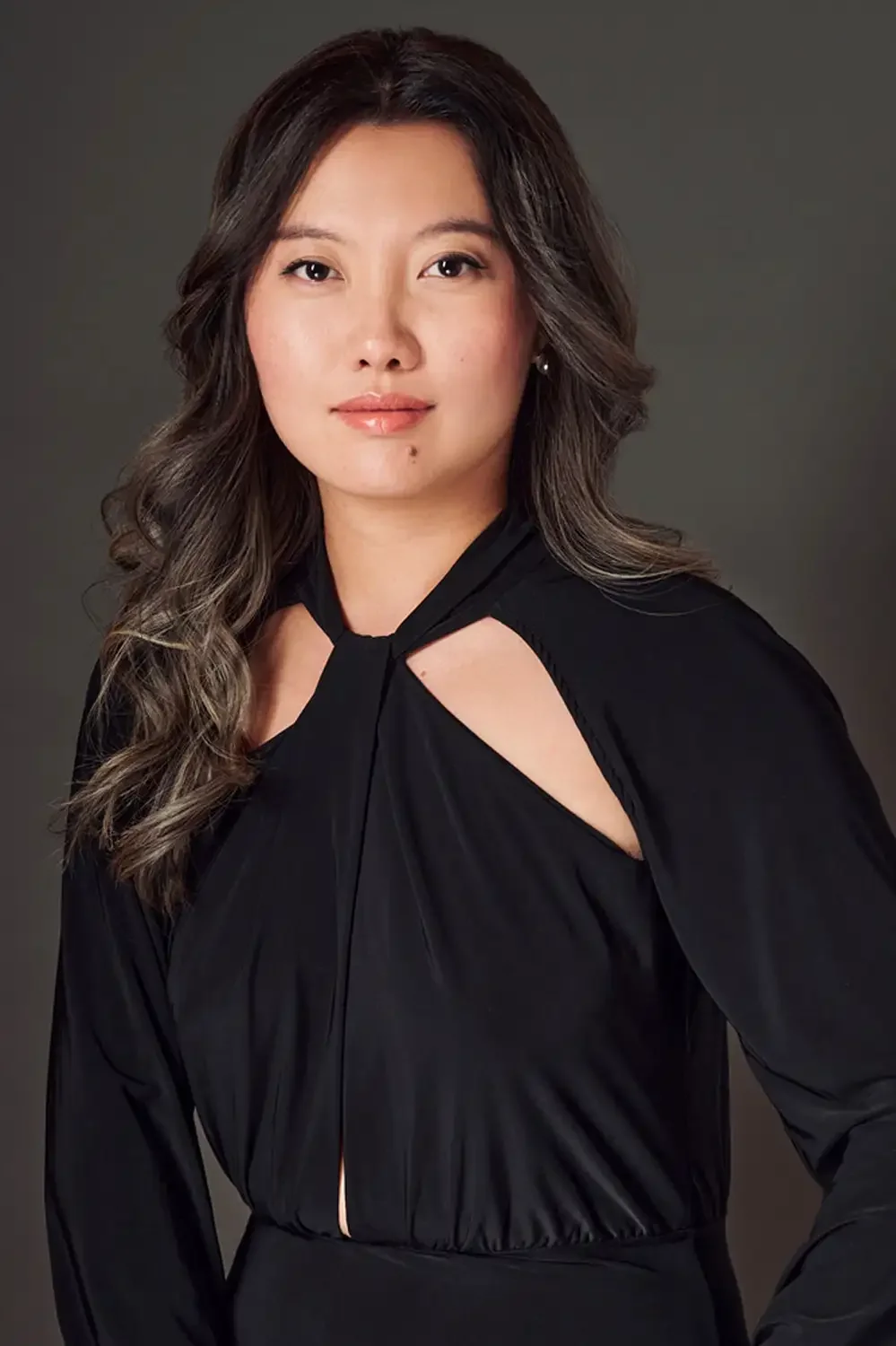 Headshot of an asian woman with wavy, shoulder-length hair wearing a black dress with unique cut-outs on the shoulders, posing in a photo studio.