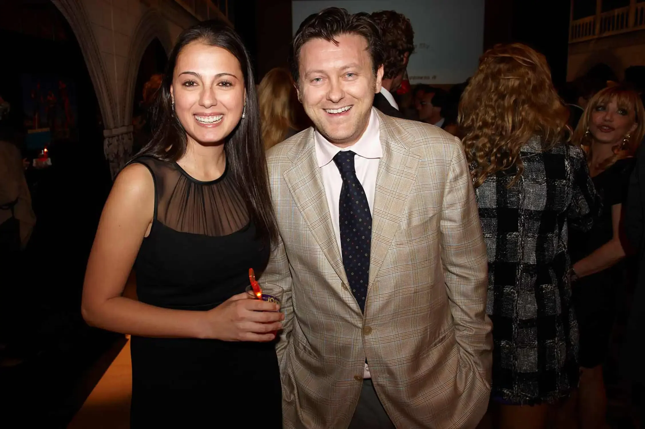 A woman with long dark hair smiling and wearing a black dress, standing next to a man in a beige suit with a white shirt and black tie, smiling at a social event.