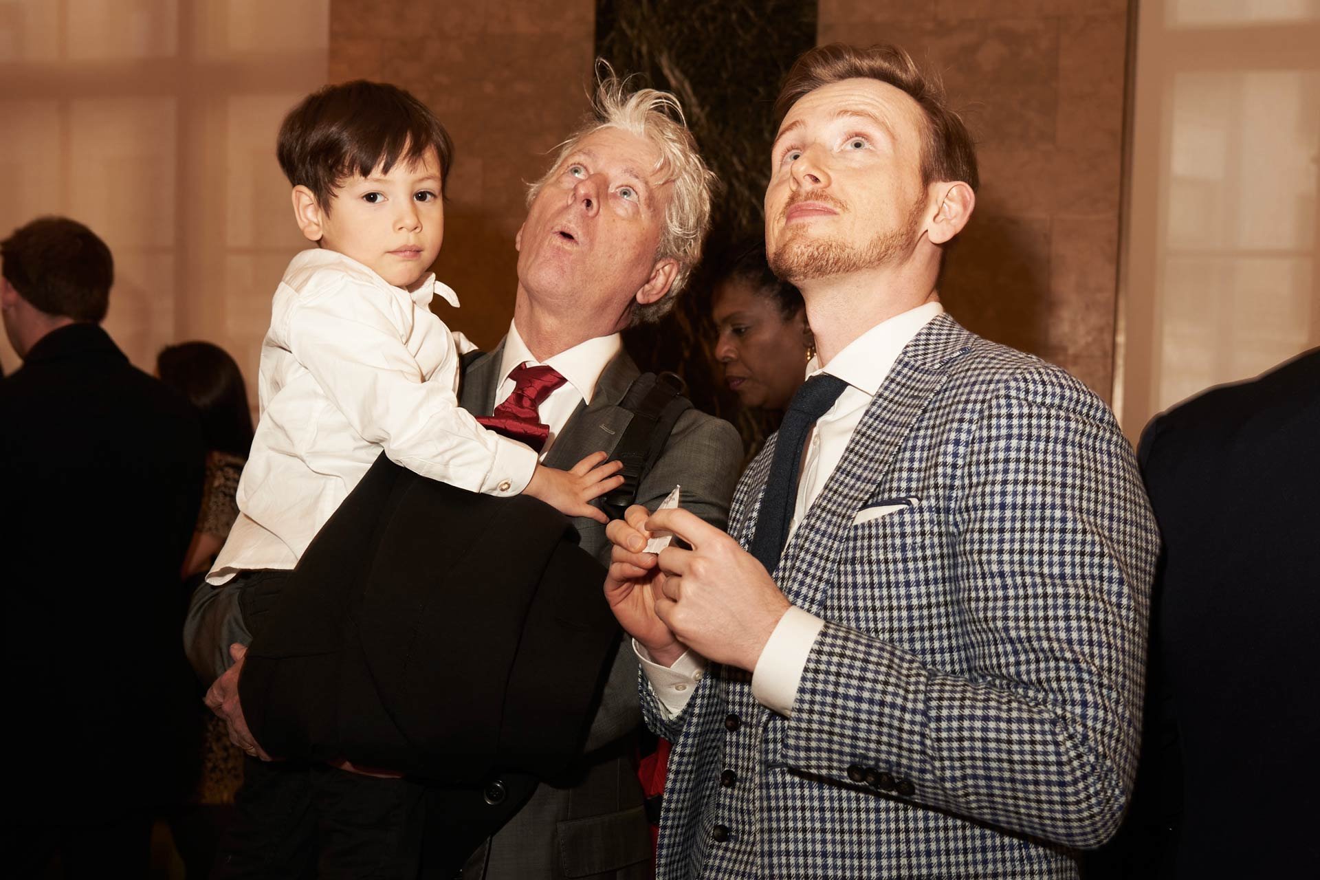 Three men and a young boy at an indoor event, with the boy sitting on one man's arm. All are looking up, with two men holding something in their hands.