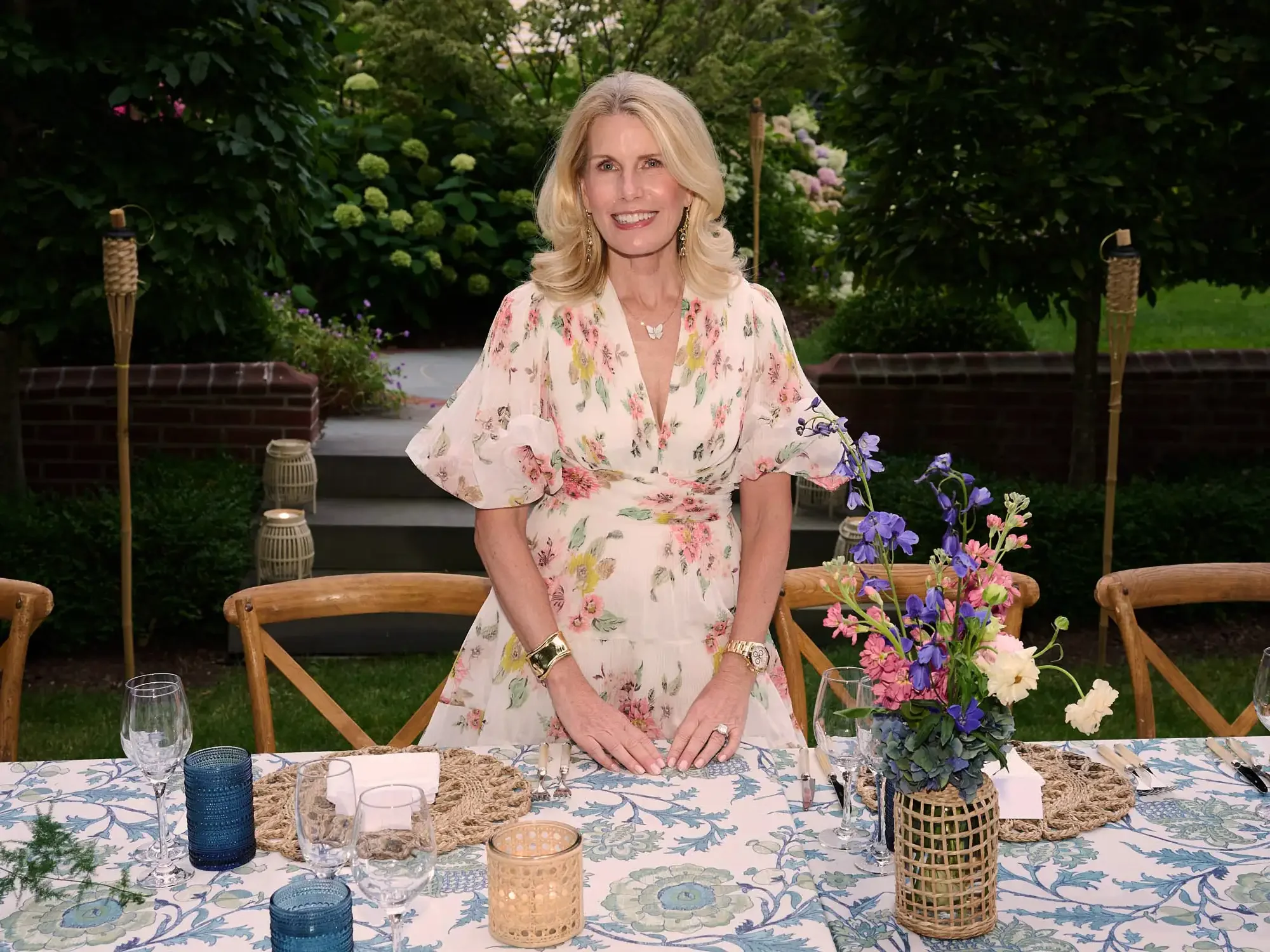 A smiling woman with blonde hair wearing a floral dress, standing at an outdoor dinner table decorated with flowers and candles, surrounded by greenery and tiki torches.
