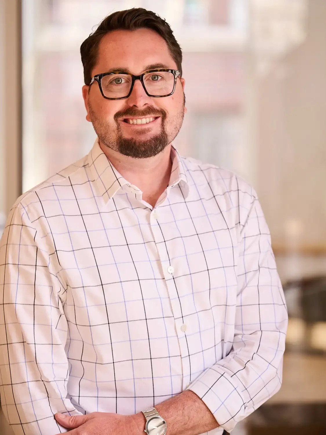 Portrait of an executive with glasses, a beard, and a mustache smiling, wearing a white shirt with a black checkered pattern, standing in office background.