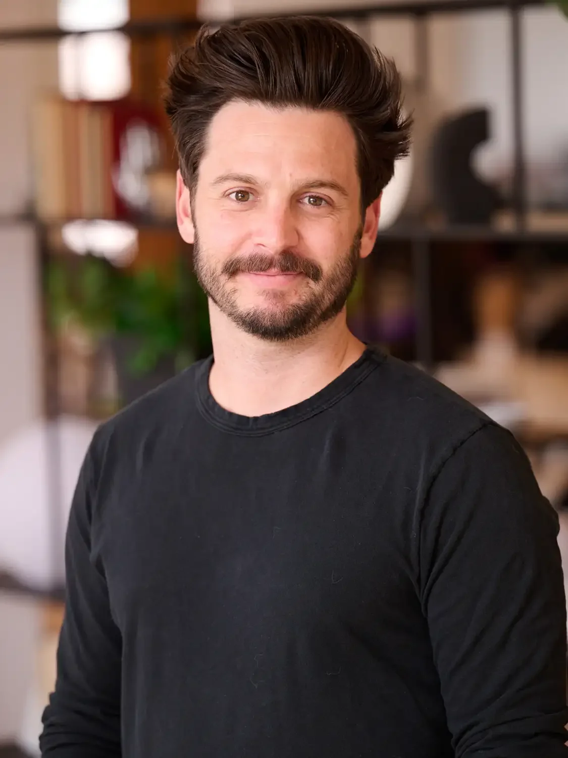 Headshot of a man with dark hair and a beard, wearing a black shirt, smiling at the camera in an office setting New-York