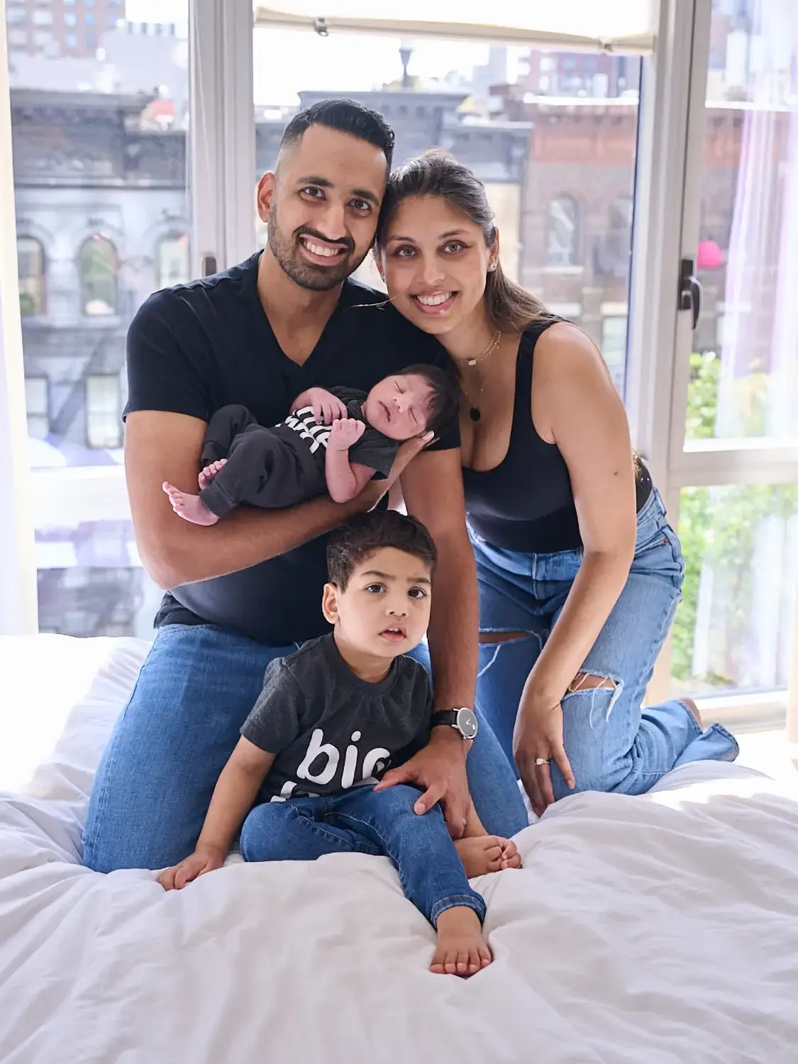 A happy family of four photographed in their apartment, in a bright room near a large window with city buildings outside. The father holds a newborn baby, and the older son sits on a bed in front of them.