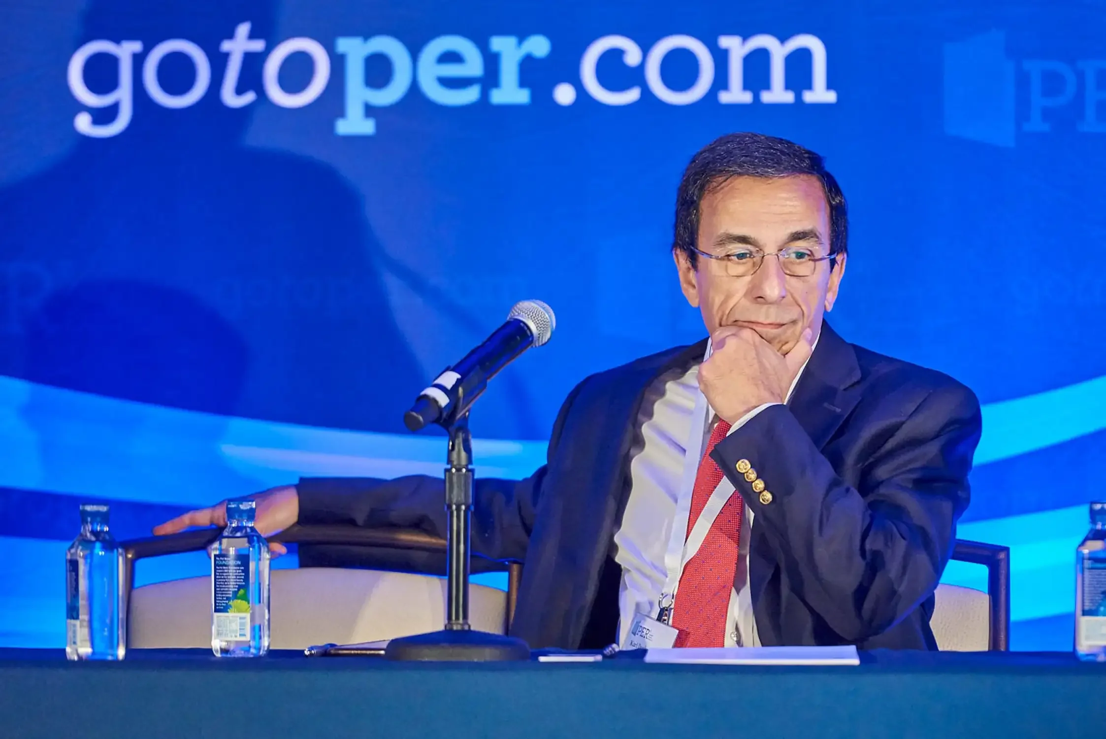 Man in a suit with glasses sitting at a conference table with a microphone, water bottles, and a name badge, in front of a blue background, event photography