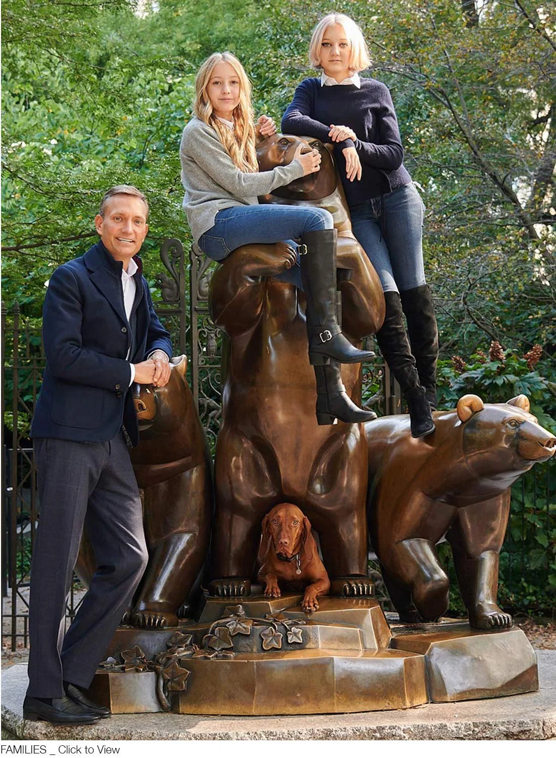 Two young girls and an adult man posing around a large bronze sculpture of bears and checkers in a park. One girl is sitting on top of the sculpture while the other is standing beside it. The man is standing with one hand on a bear's head.