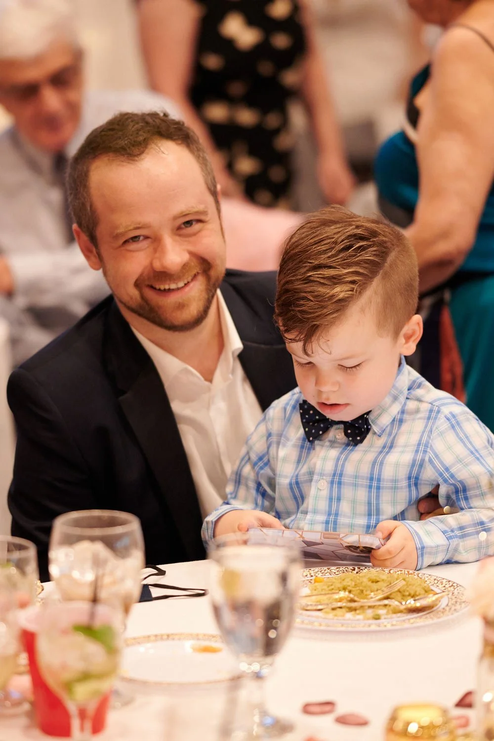 A man with a beard and a white shirt under a black blazer smiling at a camera while a young boy with a checkered shirt and a bow tie is looking down at a smartphone at a dinner table.