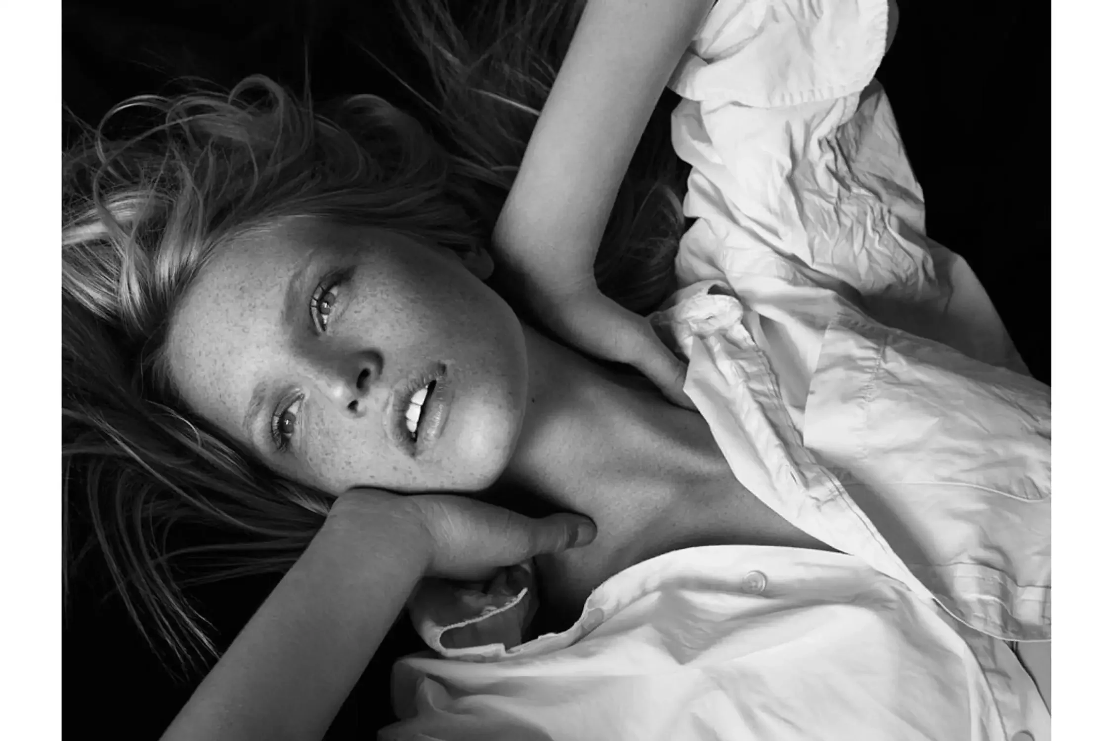 Black and white photo of a woman with freckles smiling with her eyes closed, resting her head on her hand, with long wavy hair and wearing a white shirt.