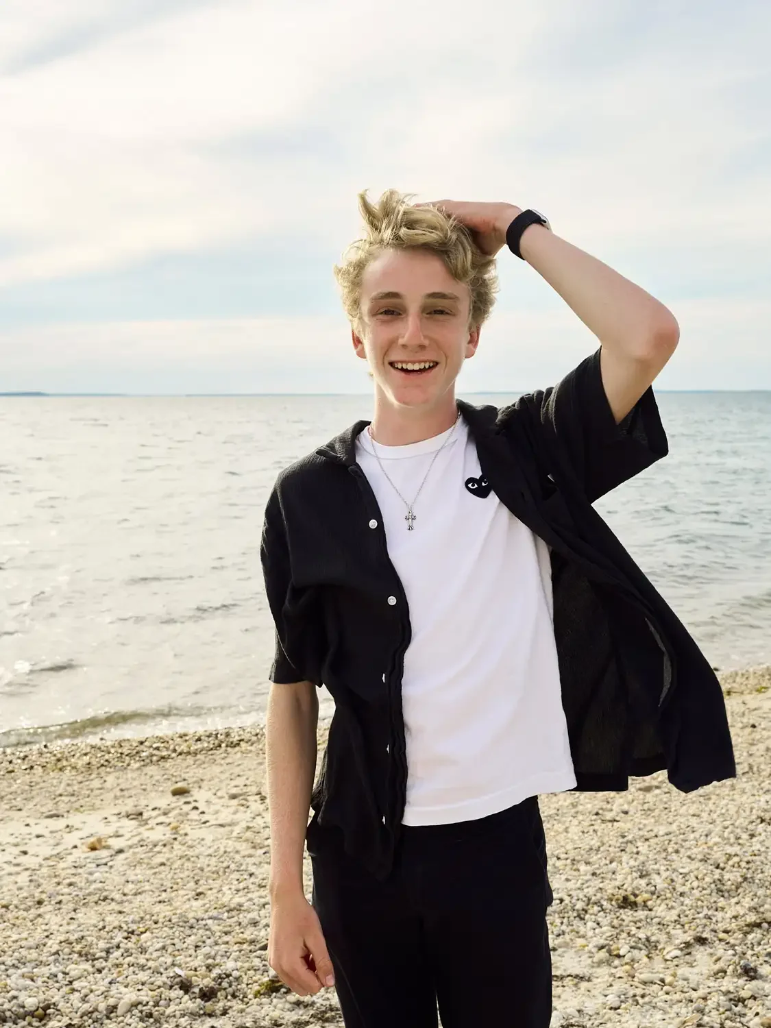 A young man smiling at the camera while standing on a beach with the ocean in the background.