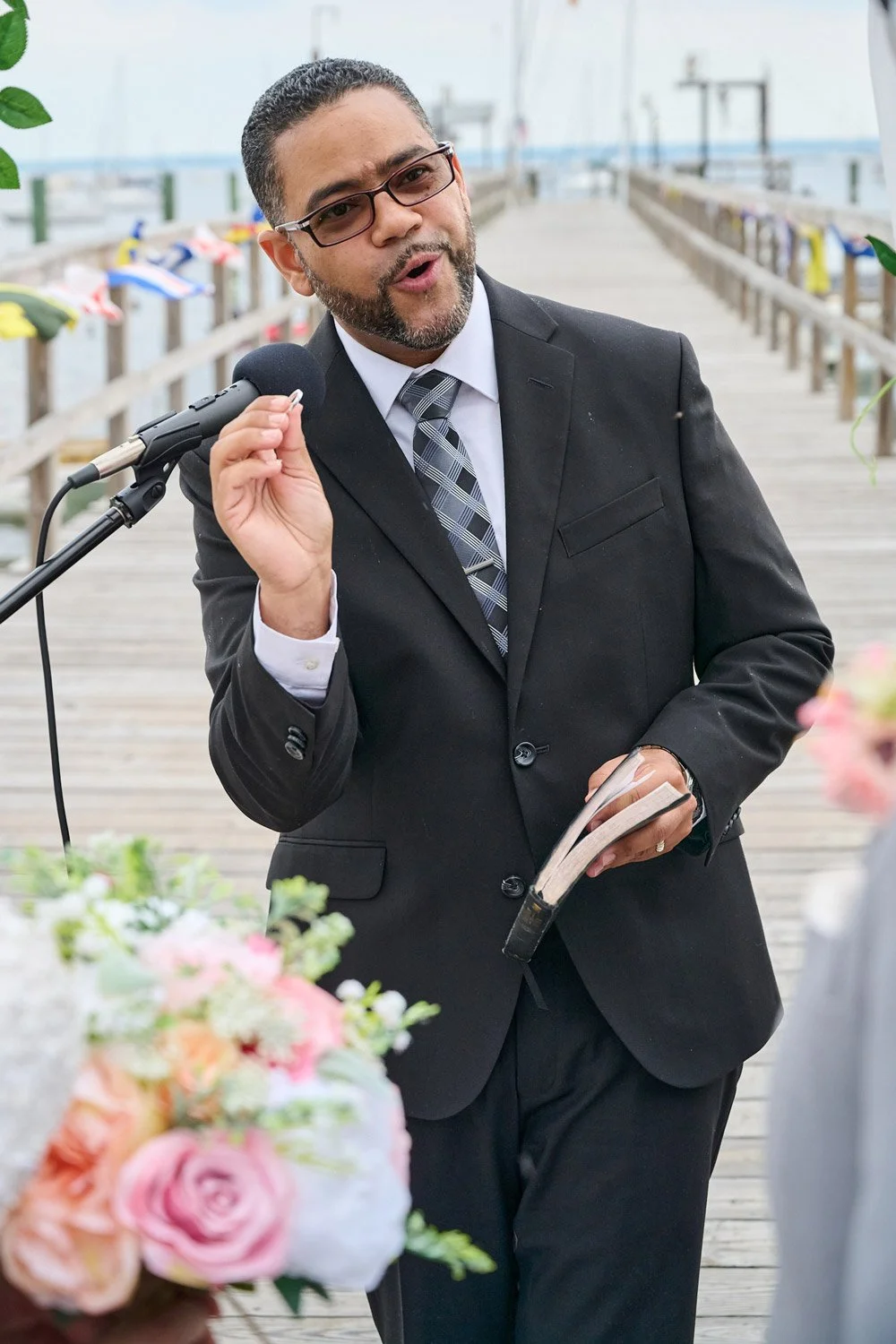 A man in a suit, holding a book, speaking into a microphone outdoors near a wooden pier with colorful umbrellas in the background.