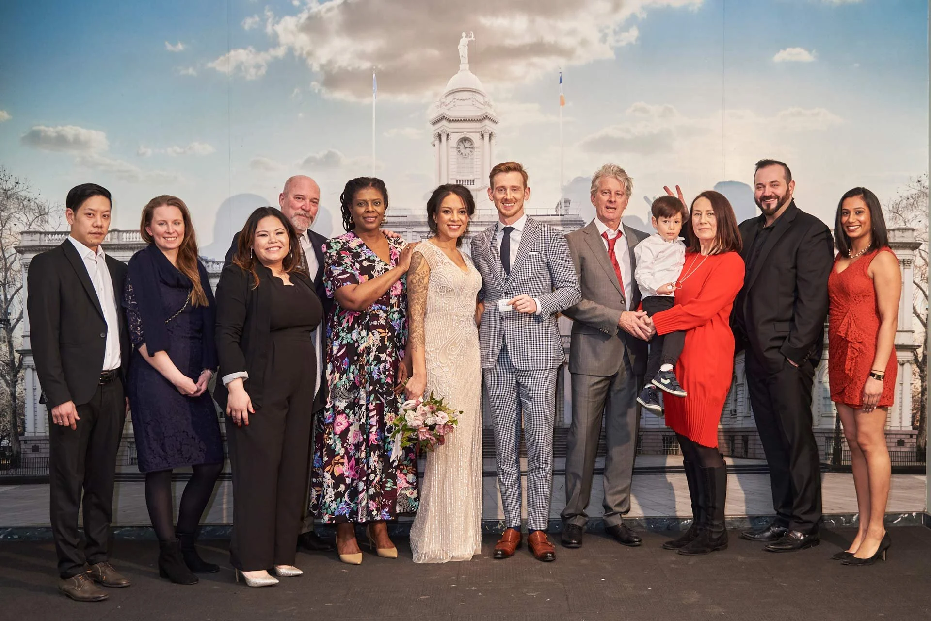 A large group of people dressed in formal attire posing for a photo in front of a backdrop of a historic building with a clock tower and a statue.