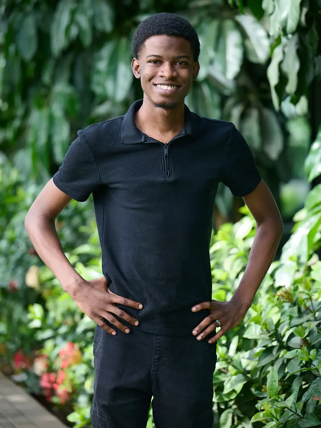 Photo of a  young man with dark skin and short curly hair, smiling and standing outdoors with lush green foliage in the background.