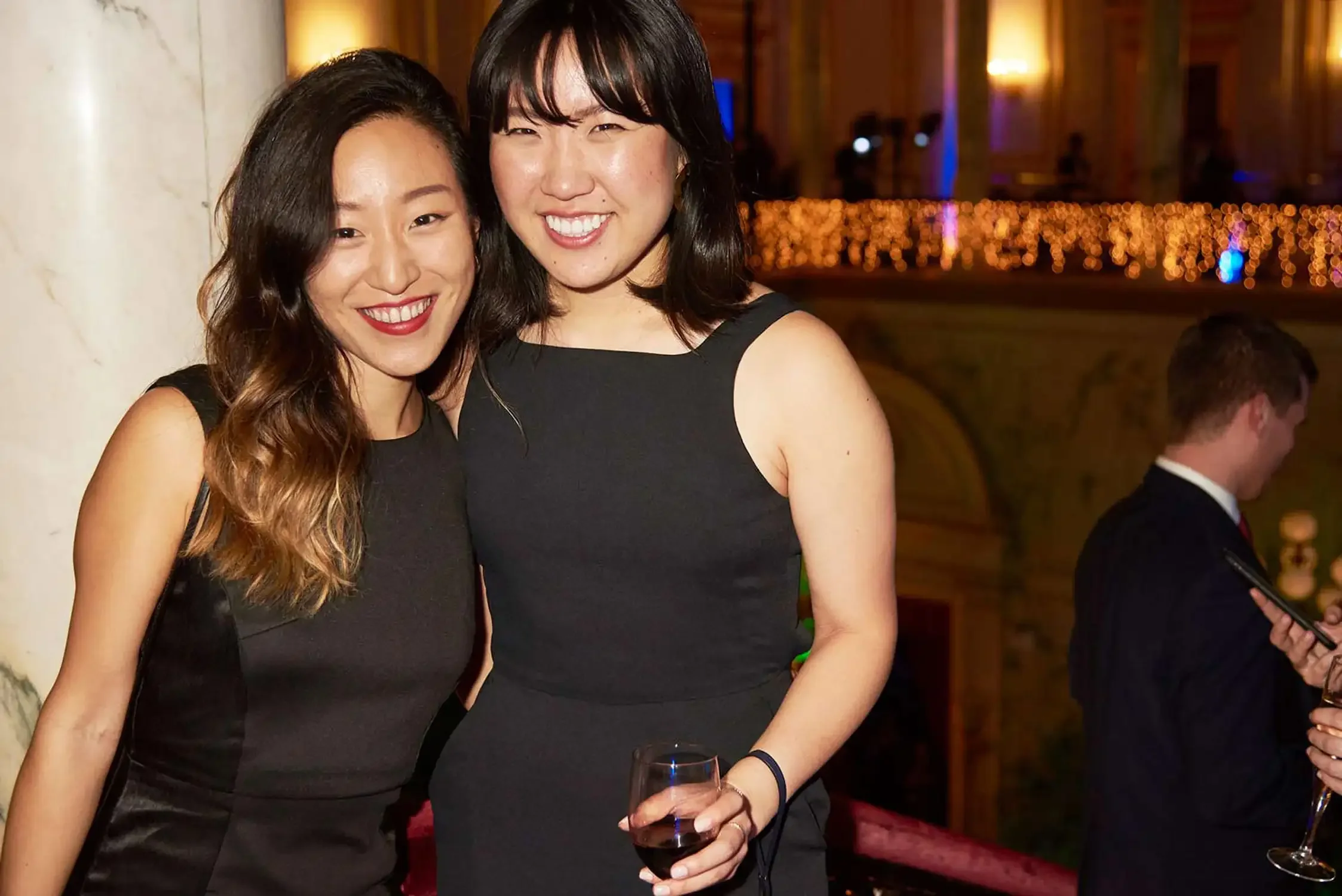 Two asian women wearing black dresses smiling at an event, one holding a glass of red wine, in an elegant venue.