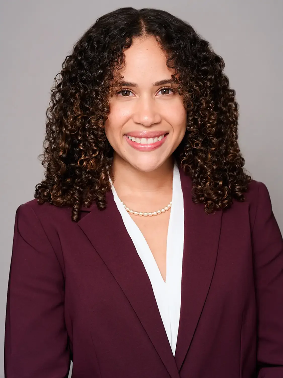 A woman with medium skin tone and curly hair smiling in a maroon blazer and pearl formal headshot in a photo studio