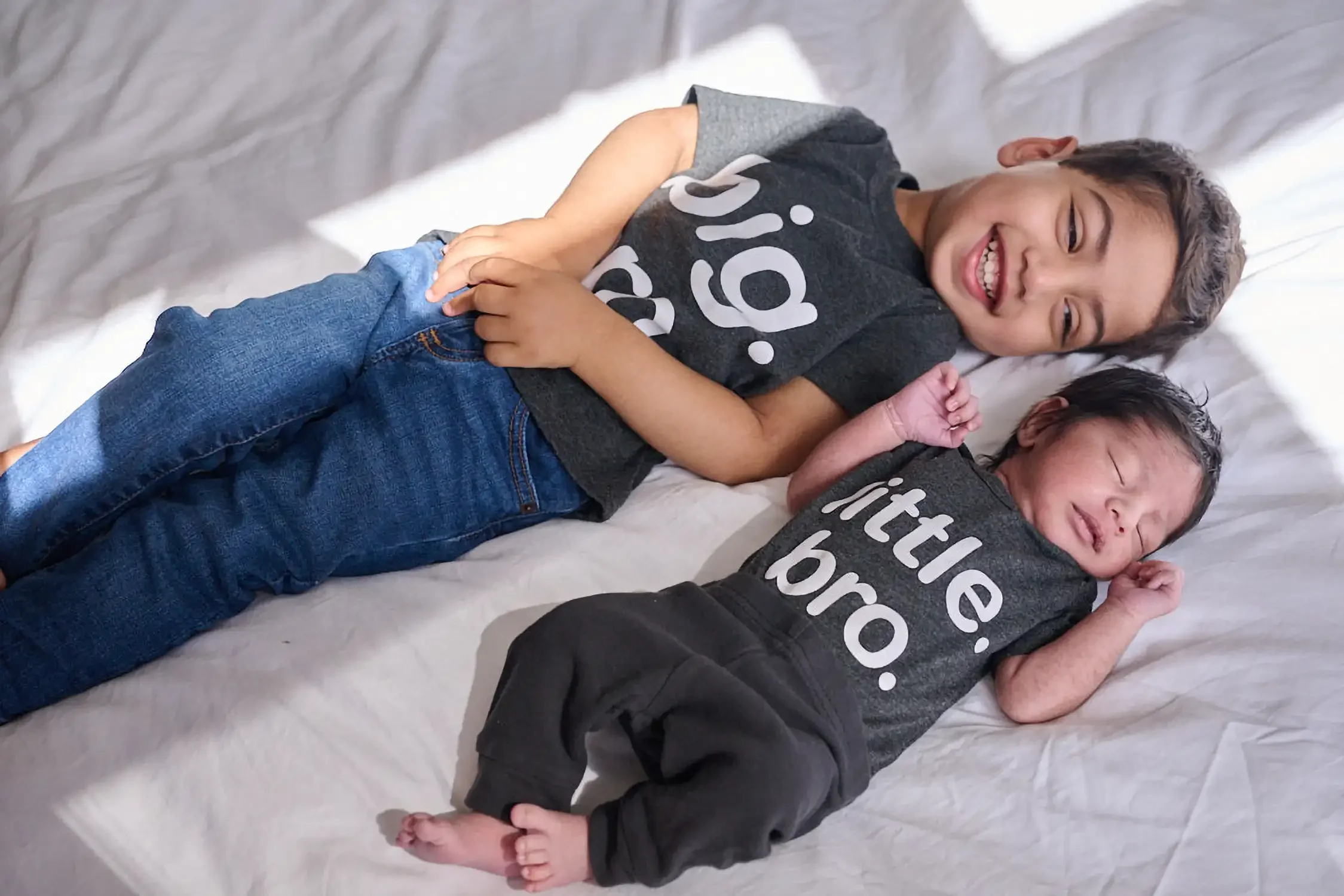 Two young children lying on a bed, smiling and touching each other, wearing matching black t-shirts with white text, and denim jeans home setting.