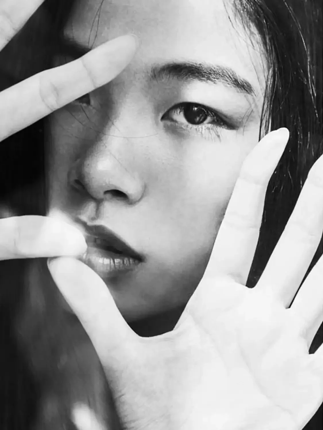 Close-up black and white photo of an Asian model's face with hands framing her face behind a window in. beautiful lighting setup.