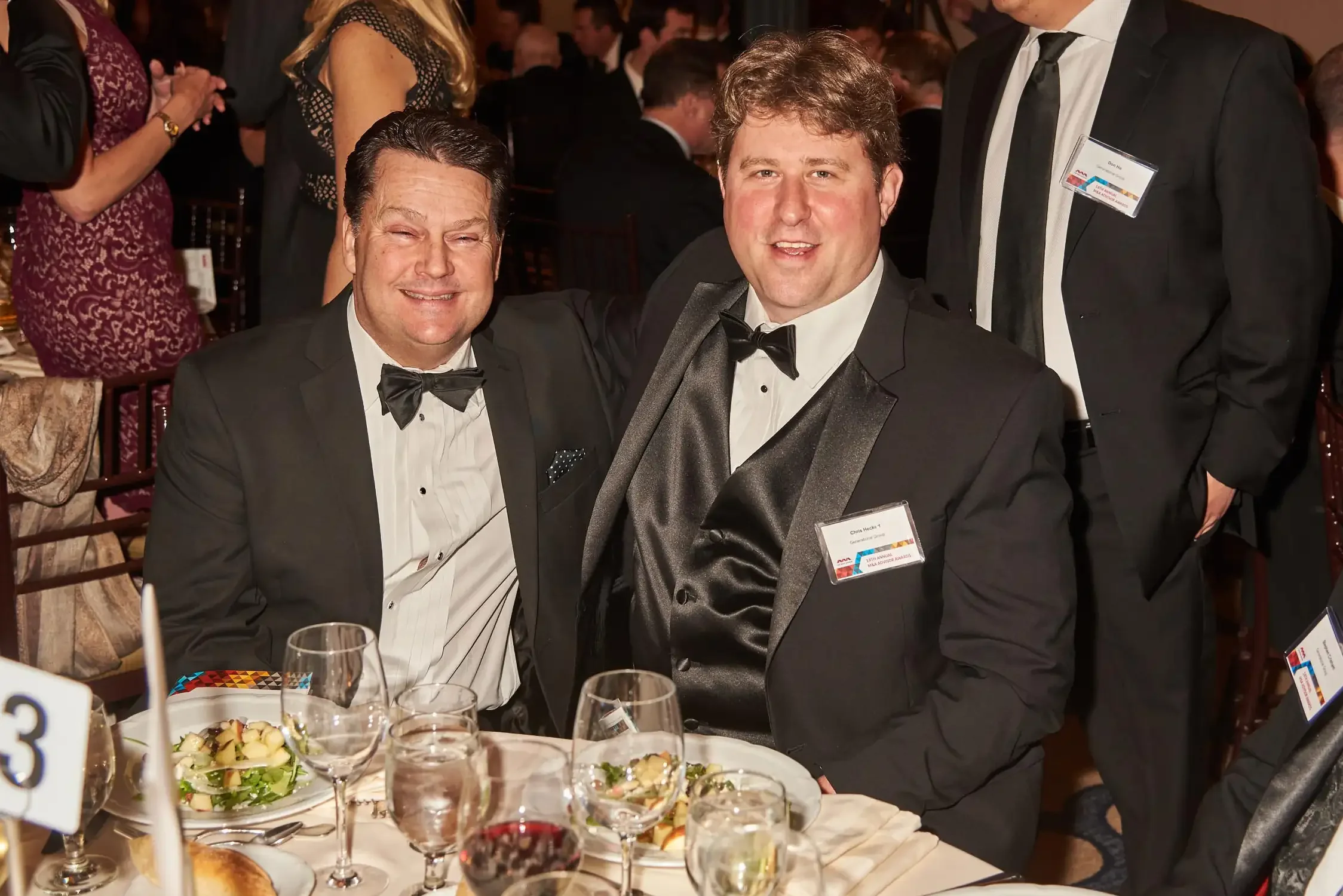 Two men in tuxedos posing together at a corporate event, seated at a table with salads, glasses, and table number cards, surrounded by other guests in formal attire.